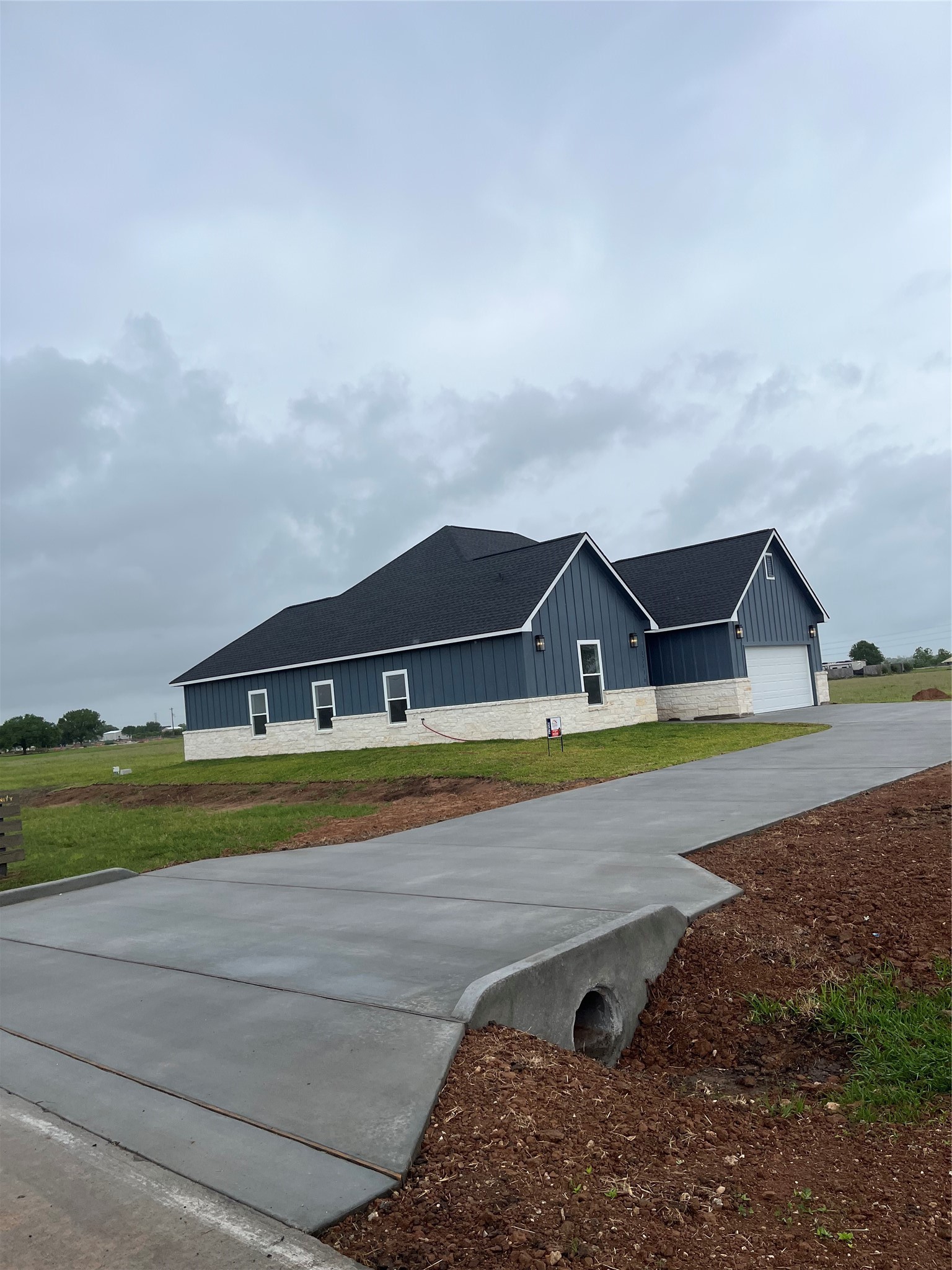 This photo features a modern, single-story home with a dark roof and blue siding, accented by white stone. It has a large driveway and is set against a backdrop of a spacious, grassy landscape under a cloudy sky.