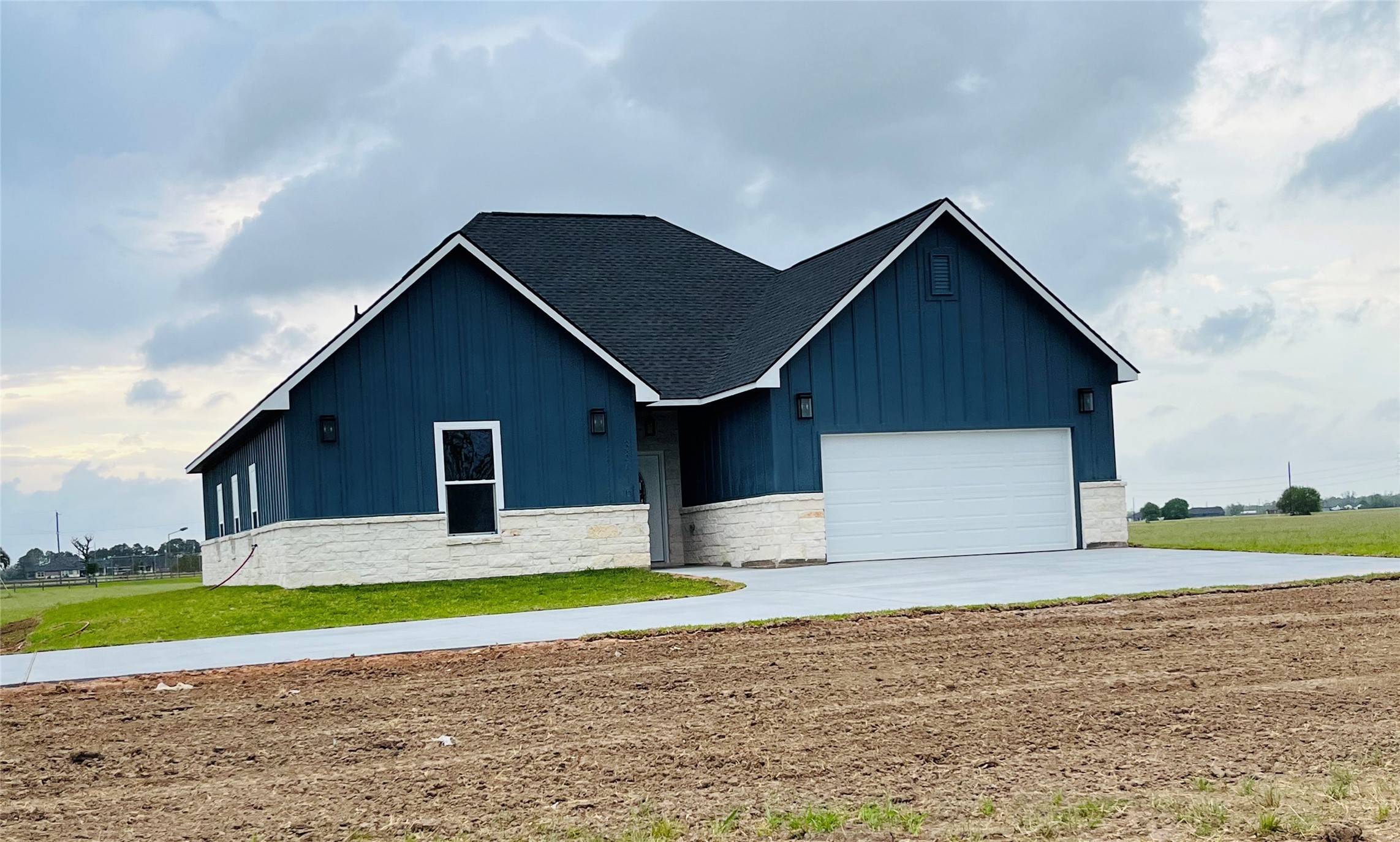 33416 FM 521 Road Angleton, TX 77515 - Photo 2 of 20 This photo shows a modern, single-story home with a blue exterior and stone accents. It features a dark roof, a two-car garage, and a spacious driveway. The house is set on a large plot with open grassy areas, providing a serene, rural feel.