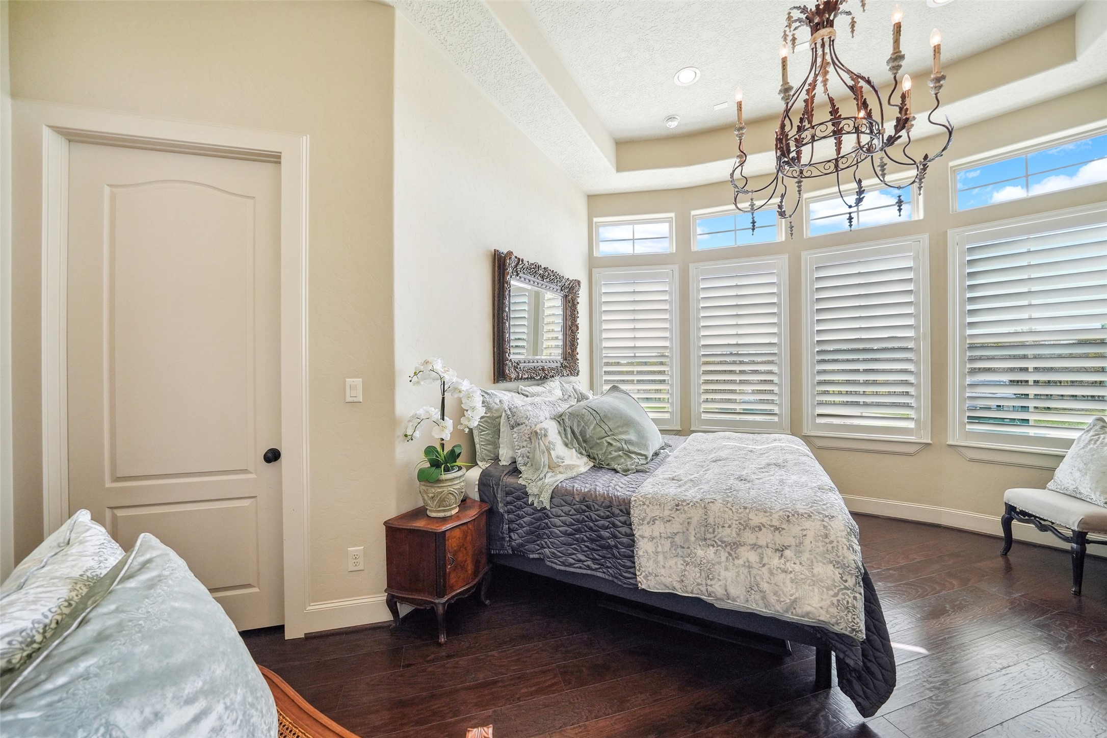 460 Bishop Road Huntsville, TX 77320 - Photo 15 of 50 A cozy bedroom featuring a curved wall of large windows fitted with custom plantation shutters, allowing for ample natural light. The ceiling showcases a unique dome design with a recessed section, adding depth and architectural interest, complemented by an elegant chandelier.