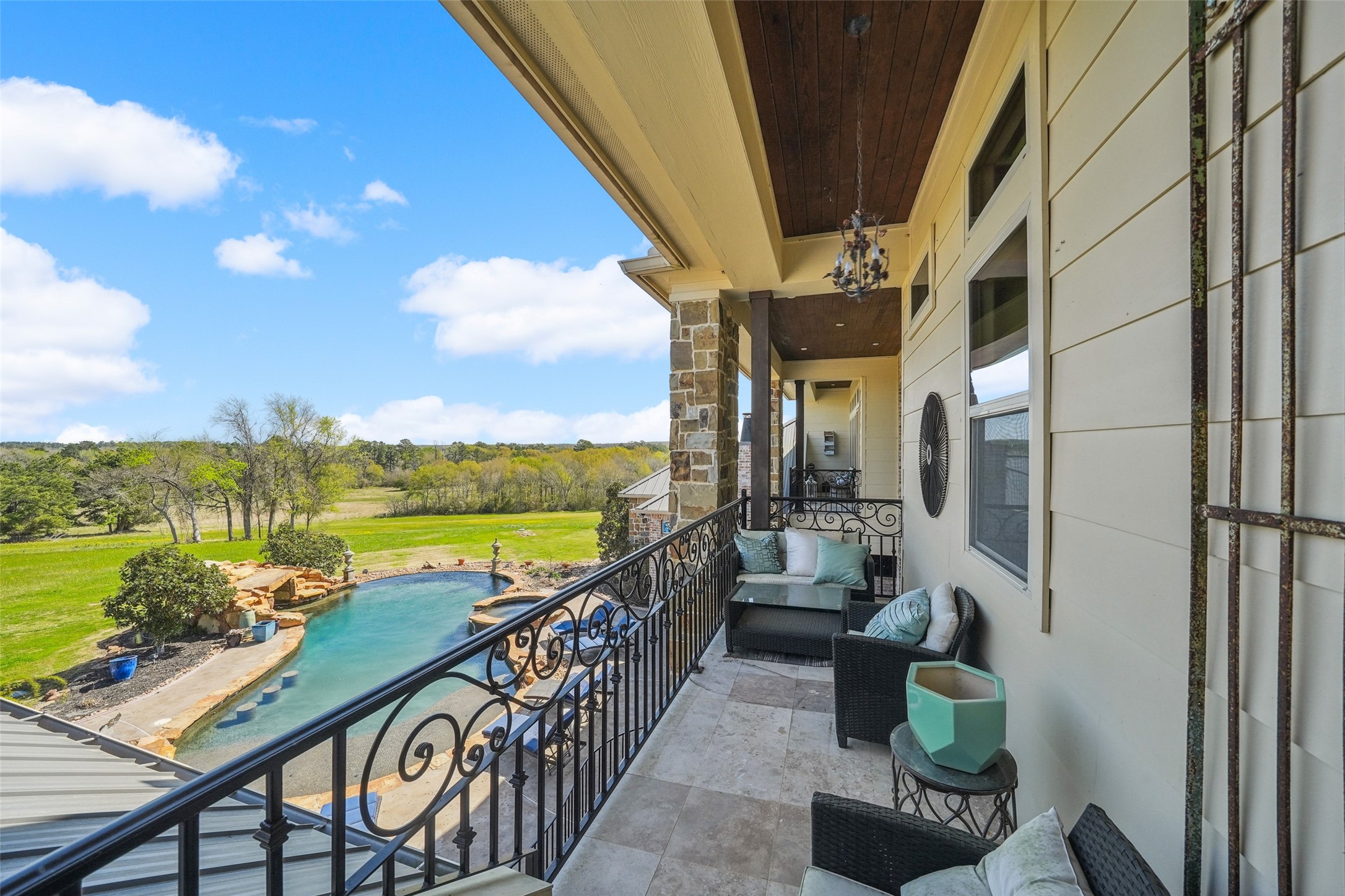 460 Bishop Road Huntsville, TX 77320 - Photo 27 of 50 A spacious balcony with a wrought iron railing, covered by a wooden ceiling with recessed lighting. The area is floored with large, light-colored tiles, and there are multiple windows along the exterior wall. The balcony overlooks a landscape with a pool and open greenery.