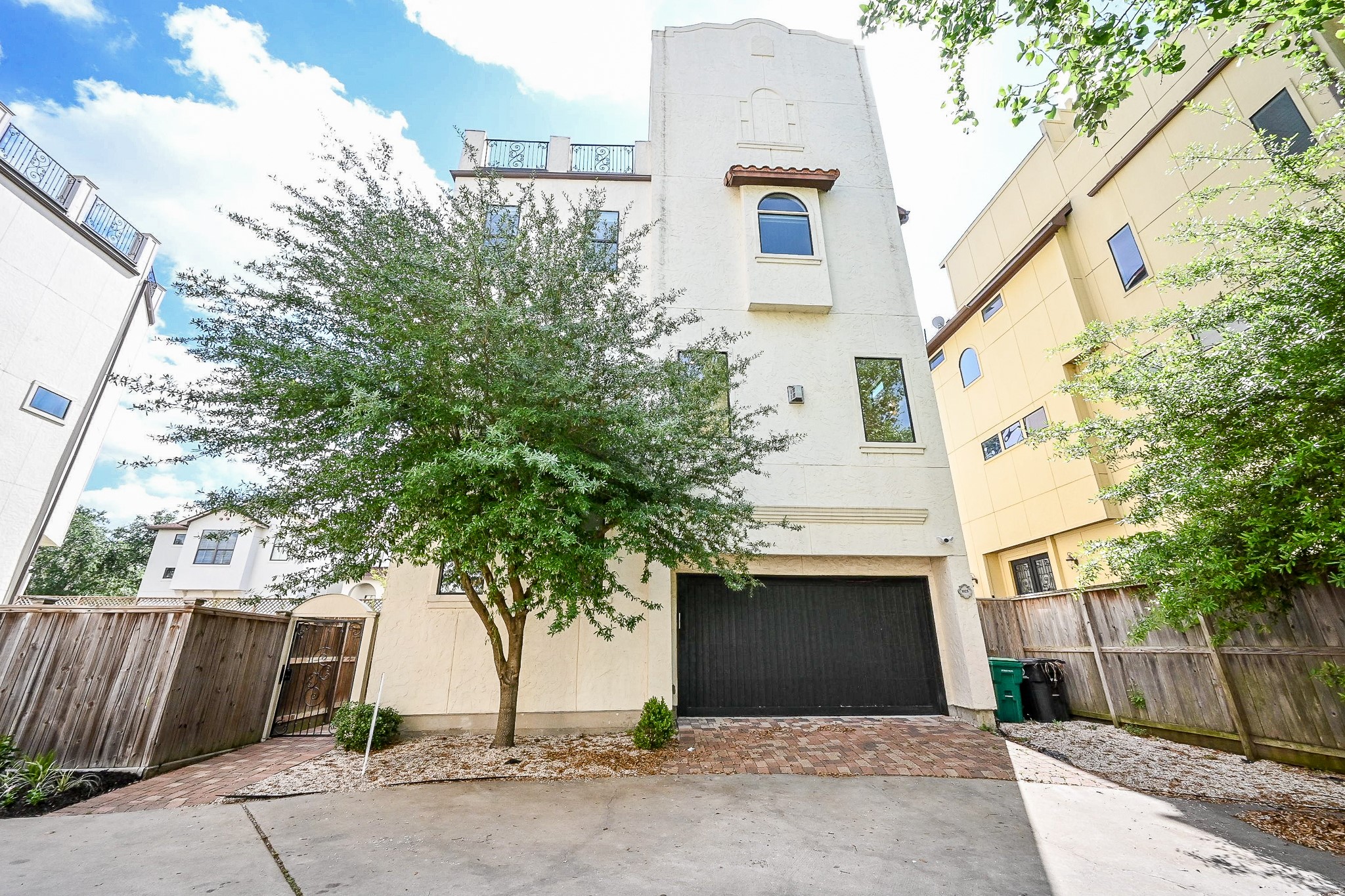 1825 West 24th Street Houston, TX 77008 - Photo 6 of 32 Modern four-story townhouse with a stucco exterior, featuring a two-car garage with an EV charger and a private fenced yard. The property is surrounded by trees, offering a blend of urban living and natural privacy.