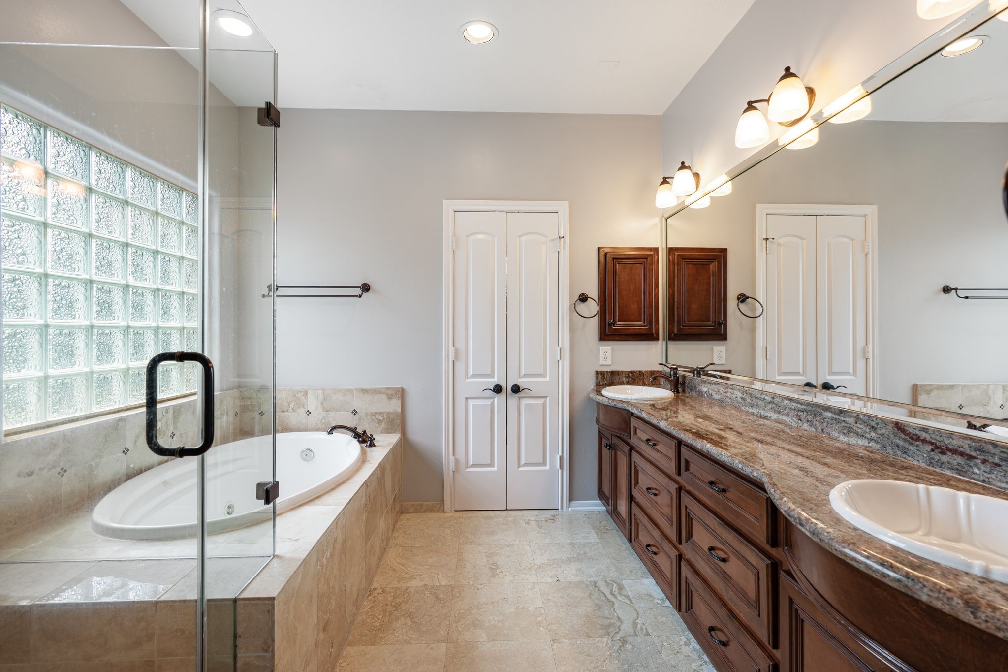 3822 Merrick Street Houston, TX 77025 - Photo 19 of 34 The primary bath features a double vanity with dark cherry cabinetry, granite countertops, dual undermount sinks, oil-rubbed bronze fixtures, and a full-width mirror with multi-bulb sconce lighting above. A frameless glass shower enclosure and a jetted soaking tub surrounded by travertine tile sit alongside a glass block window that brings in natural light. Two separate closet doors provide ample storage access.