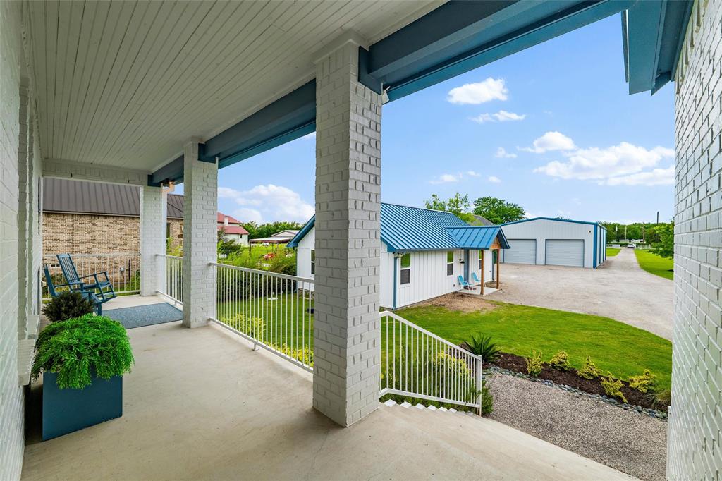 6625 Sandy Beach Road Azle, TX 76020 - Photo 6 of 40 Front porch features painted brick support columns, exposed beam accents, metal railing detail, and concrete flooring throughout the covered entry area. Exterior overlooks landscaped lawn spaces, gravel driveway access, and views toward the detached guest house and garage structures.