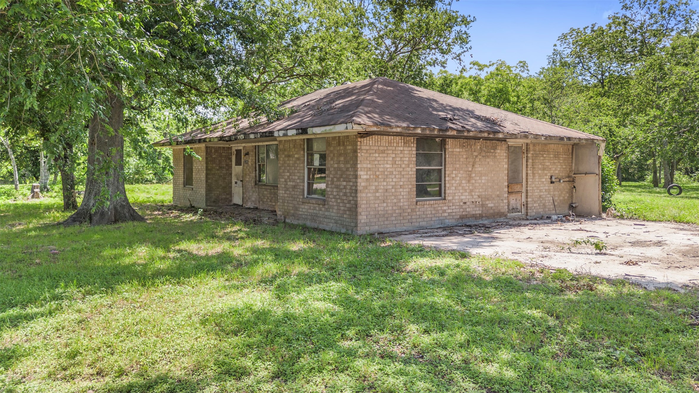 2701 Mazen Road Rosenberg, TX 77471 - Photo 3 of 10 This photo shows a single-story brick house situated on a spacious, grassy lot with mature trees. The home appears to need some renovation but is surrounded by plenty of open green space, offering a peaceful, natural setting.
