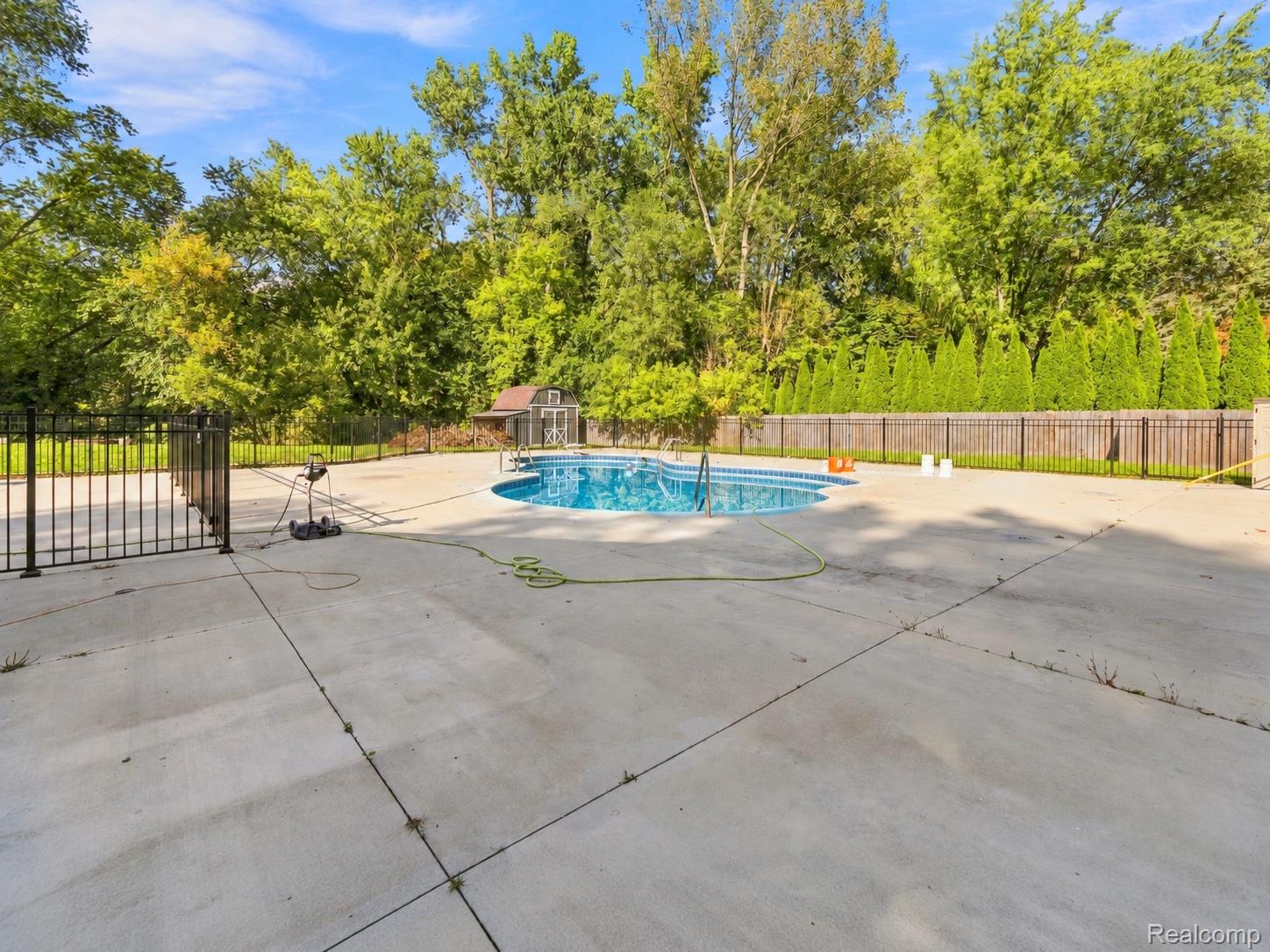 15879 Millar Road Clinton Township, MI 48036 - Photo 65 of 65 In-ground swimming pool with surrounding concrete patio, secured by a black metal fence, and bordered by mature trees