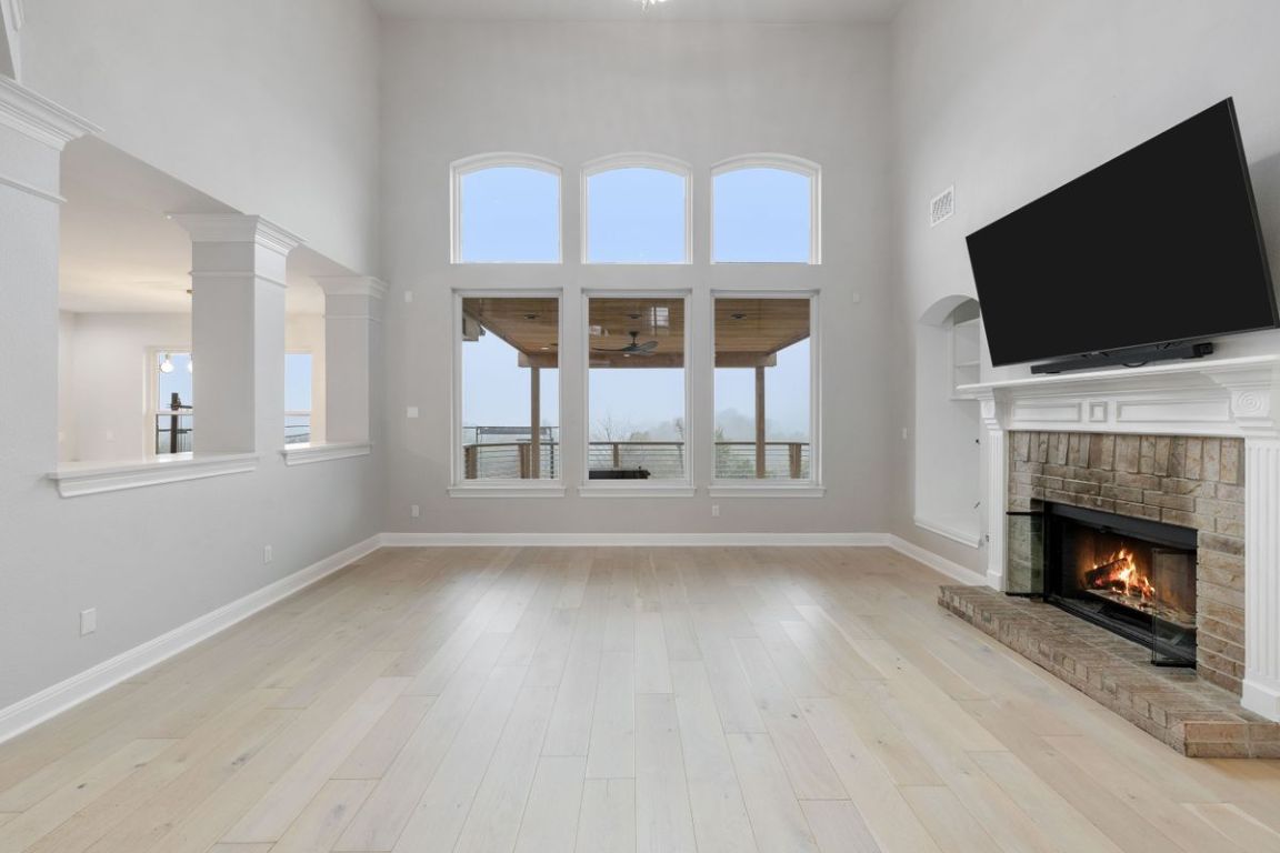 2137 Rivina Drive Austin, TX 78733 - Photo 9 of 39 Unfurnished living room featuring a brick fireplace, a high ceiling, light wood finished floors, and a ceiling fan