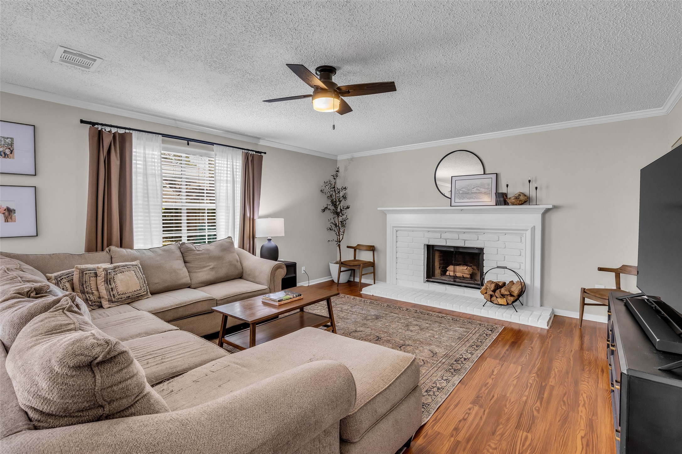 9415 Towerstone Drive Spring, TX 77379 - Photo 19 of 44 This cozy living room features a large sectional sofa, a white brick fireplace with stylish decor, and a ceiling fan. Natural light pours in through a window with elegant curtains, highlighting the wood flooring and inviting ambiance.
