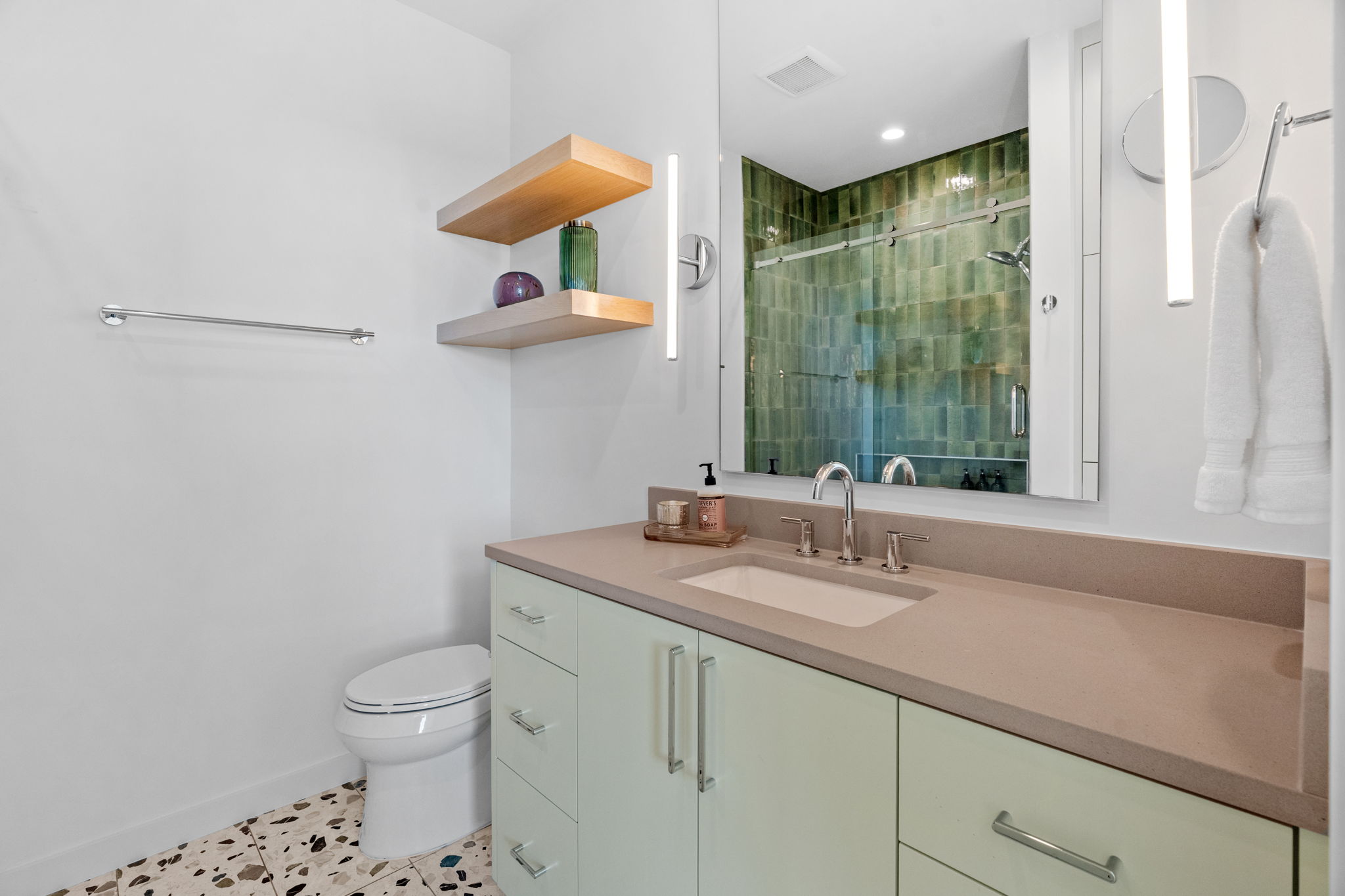 503 Happy Trail Dripping Springs, TX 78620 - Photo 26 of 38 Bathroom featuring a light green vanity with integrated sink, a large mirror with vertical sconces, a shower with green subway tile, floating wood shelving, and patterned terrazzo-style flooring
