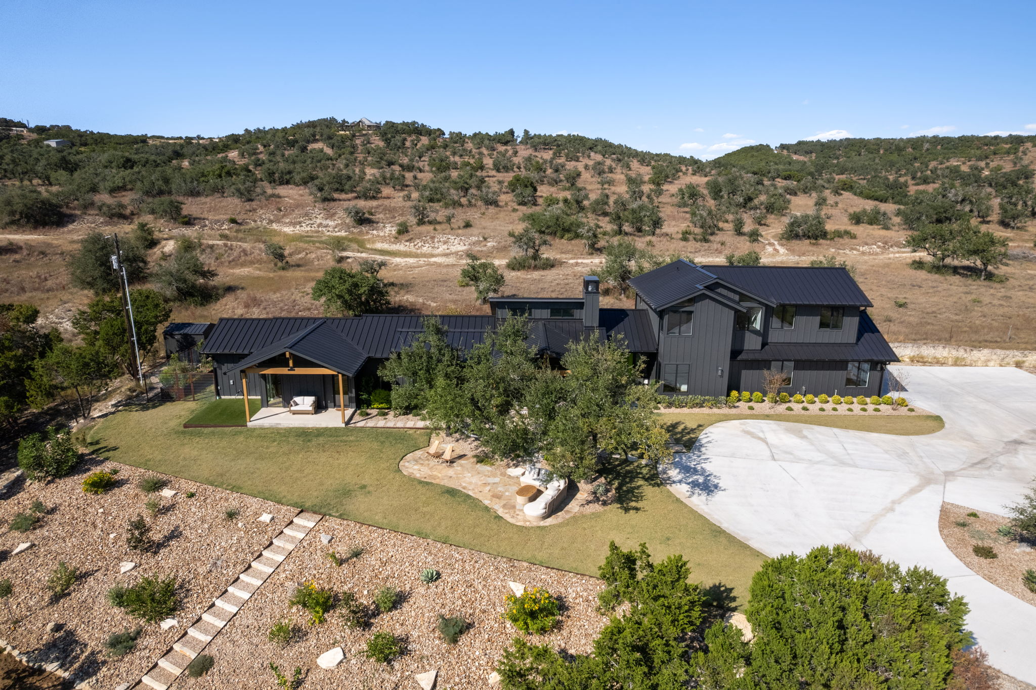 503 Happy Trail Dripping Springs, TX 78620 - Photo 38 of 38 Contemporary-style residence featuring a dark-toned exterior with a black metal roof, a covered outdoor patio, a manicured lawn, and an expansive concrete driveway