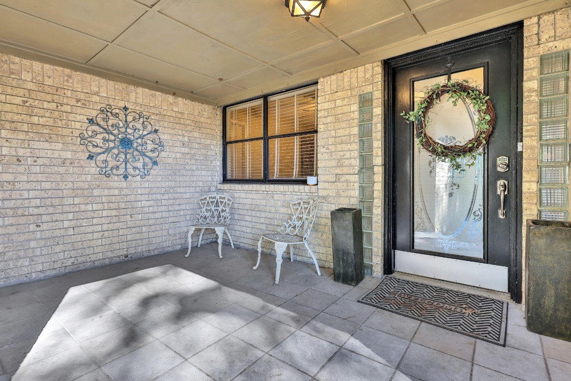 Charming front porch with tiled flooring and decorative brickwork. Features an elegant glass door with a wreath, metal wall art, and two vintage-style chairs, perfect for relaxing.