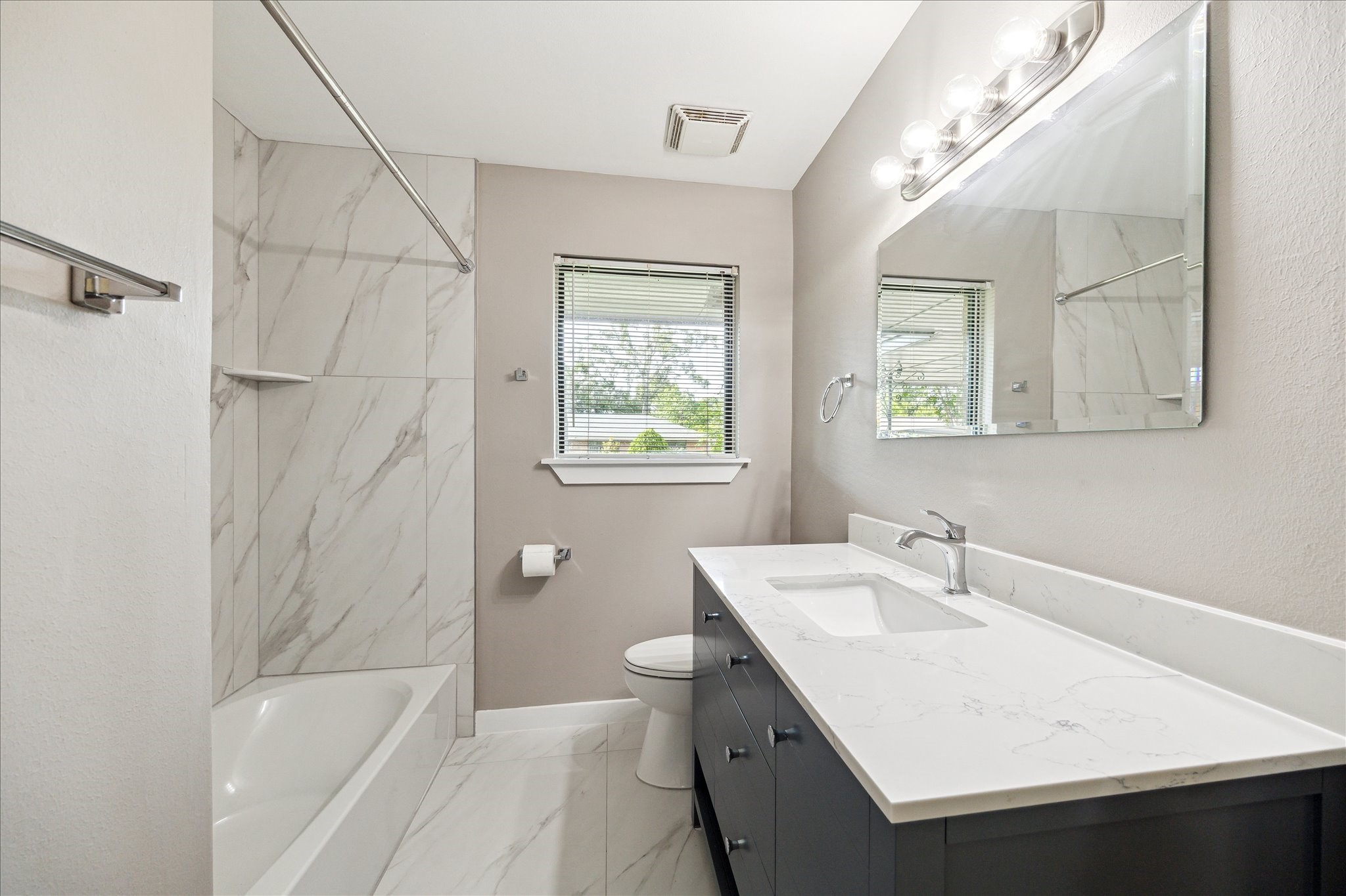 8907 Autauga Street Houston, TX 77080 - Photo 16 of 20 Modern bathroom with sleek marble tiles, a spacious vanity with a large mirror, and natural light from a window. The room includes a bathtub with a shower and elegant fixtures.