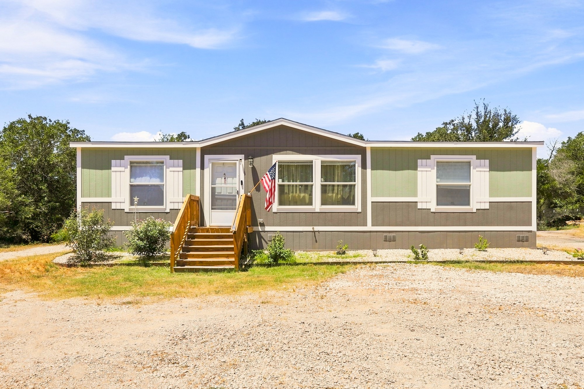 1039 Trailside Elgin, TX 78621 - Photo 4 of 24 A manufactured home with vertical siding, front steps, and a gravel driveway under clear skies.