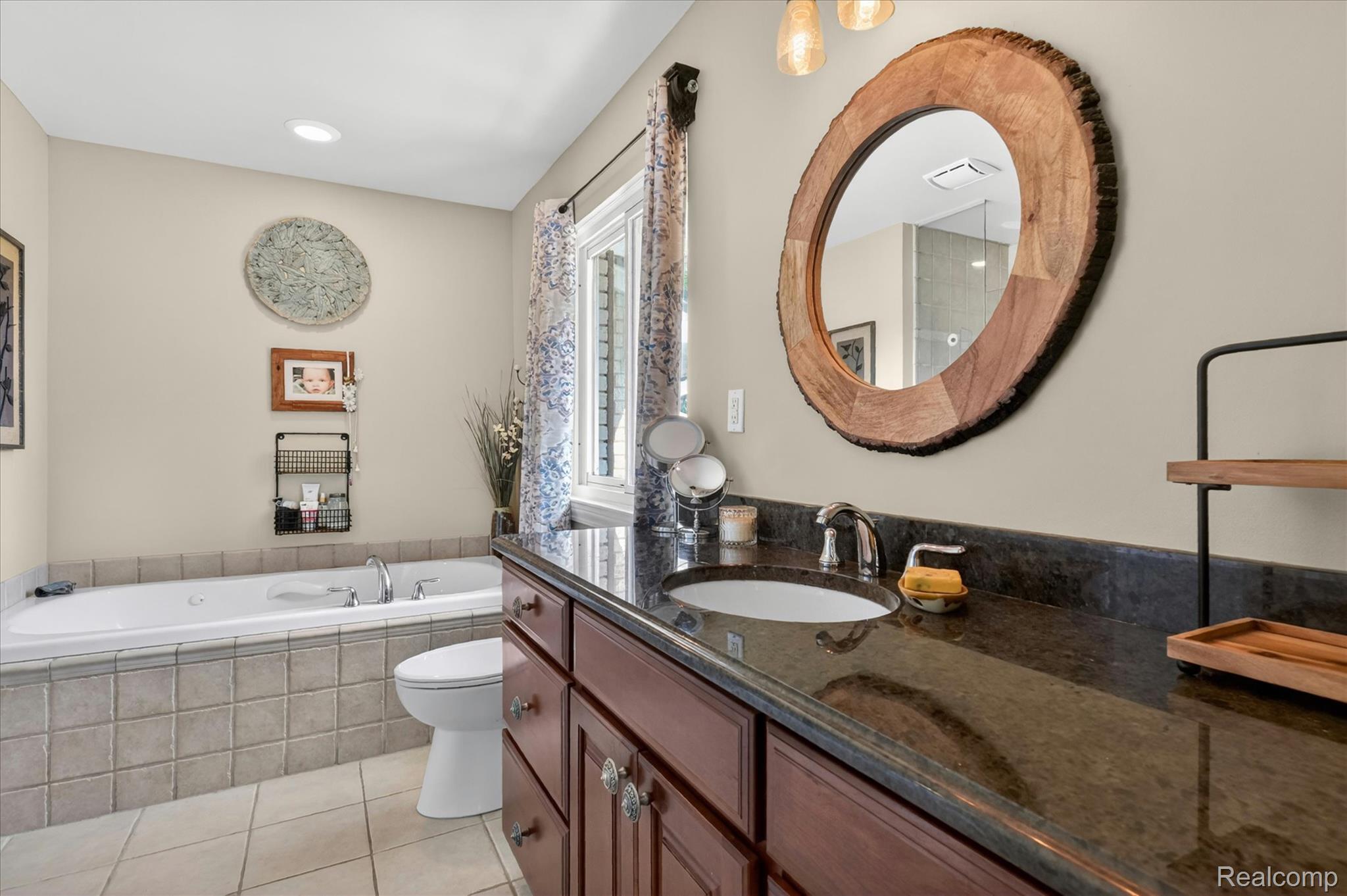 3860 East Commerce Road Commerce Township, MI 48382 - Photo 30 of 72 Bathroom featuring a dark wood-finish vanity with a built-in sink and dark countertop, a large round wood-framed mirror, a window with patterned drapes, a built-in bathtub with tiled surround, and light tile flooring