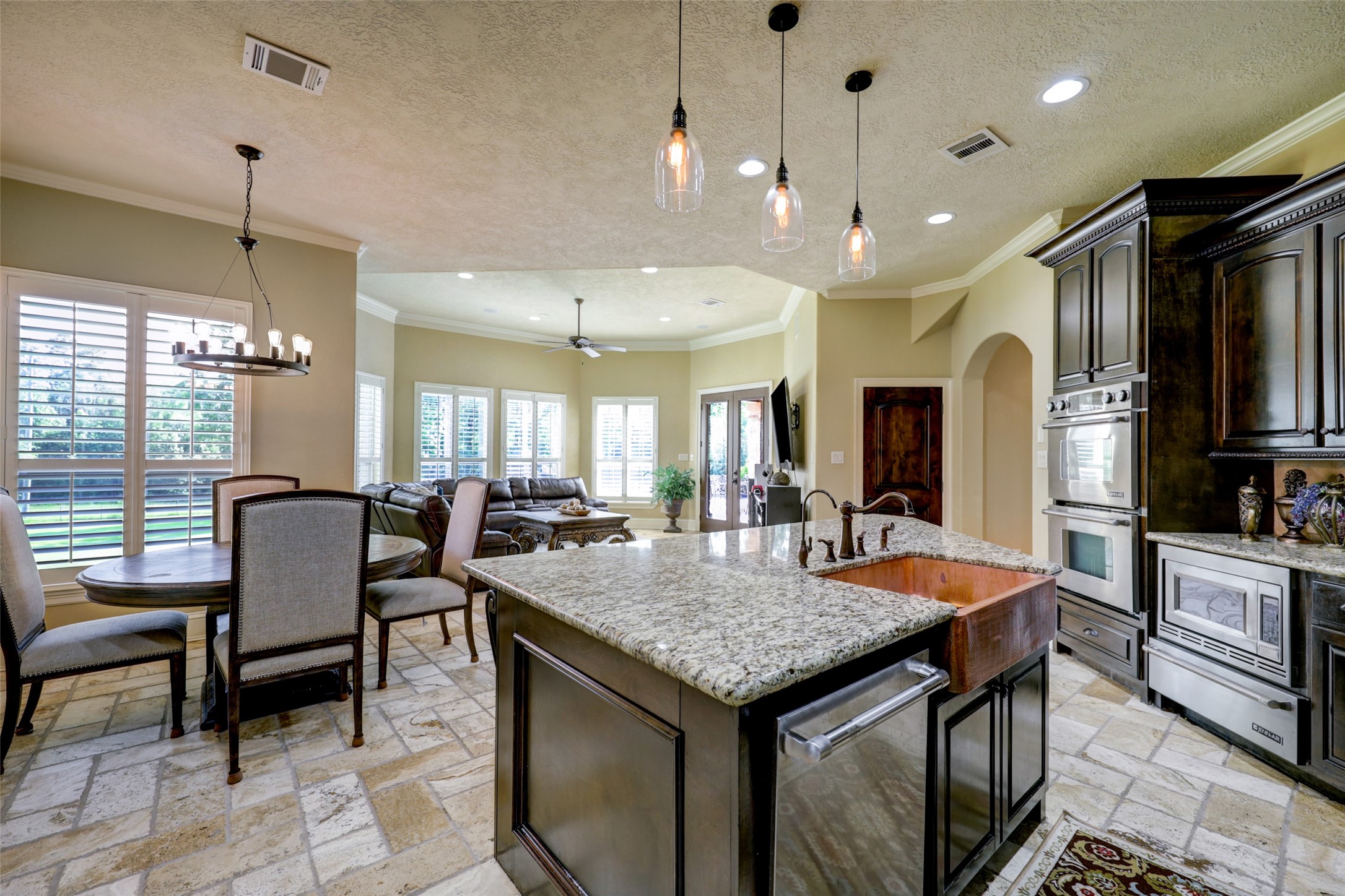 4434 Chateau Creek Way Spring, TX 77386 - Photo 16 of 34 This photo showcases a spacious open-concept kitchen and living area. The kitchen features granite countertops, a large island with a farmhouse sink, and modern appliances. Adjacent is a cozy dining area with large windows and natural light. The living space has high ceilings and access to the outdoors through glass doors.