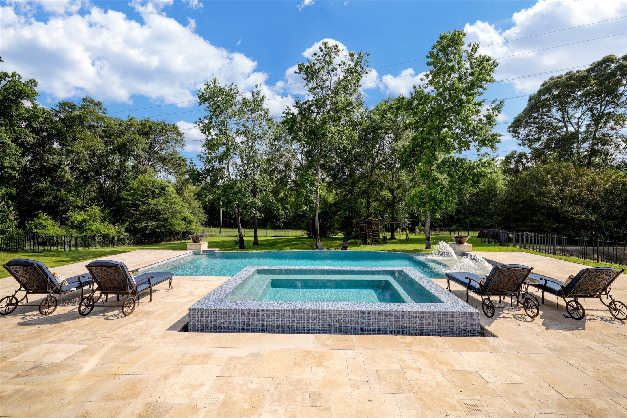 4434 Chateau Creek Way Spring, TX 77386 - Photo 2 of 34 This photo showcases a luxurious backyard featuring a modern infinity swimming pool with spa, surrounded by a spacious stone patio. The area is beautifully landscaped with lush greenery and mature trees, creating a serene and private outdoor oasis.