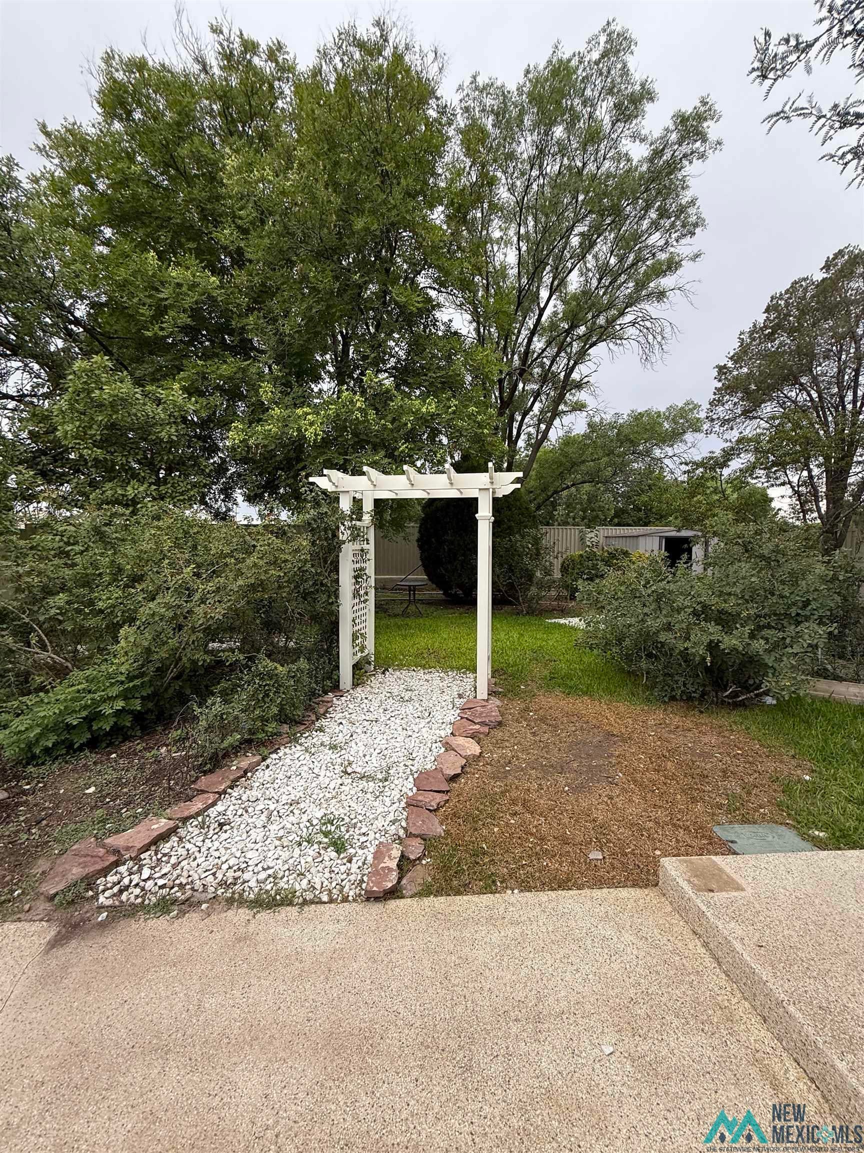 1111 West Country Road Roswell, NM 88201 - Photo 23 of 41 Backyard featuring a white pergola entryway, gravel pathway bordered by natural stone, mature trees, and a storage shed
