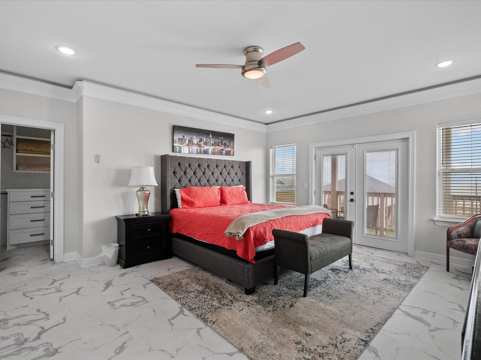 1018 Seashell Drive Surfside Beach, TX 77541 - Photo 41 of 49 Spacious bedroom with a modern design featuring a tufted headboard and red bedding. It has a ceiling fan, ample natural light from multiple windows, and French doors leading outside. The room includes a walk-in closet and elegant marble flooring.