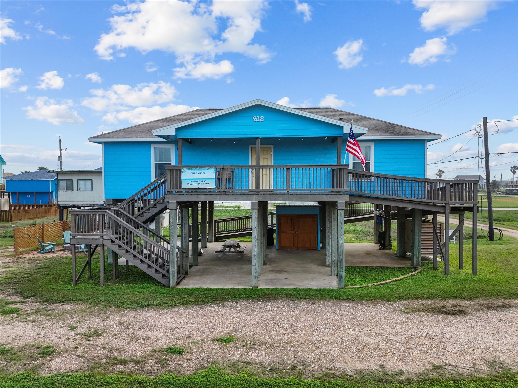 This elevated, vibrant blue home features a spacious wraparound deck and ample covered space underneath. The property offers a unique design with dual staircases and an American flag accenting the front. Perfect for those seeking a coastal vibe.