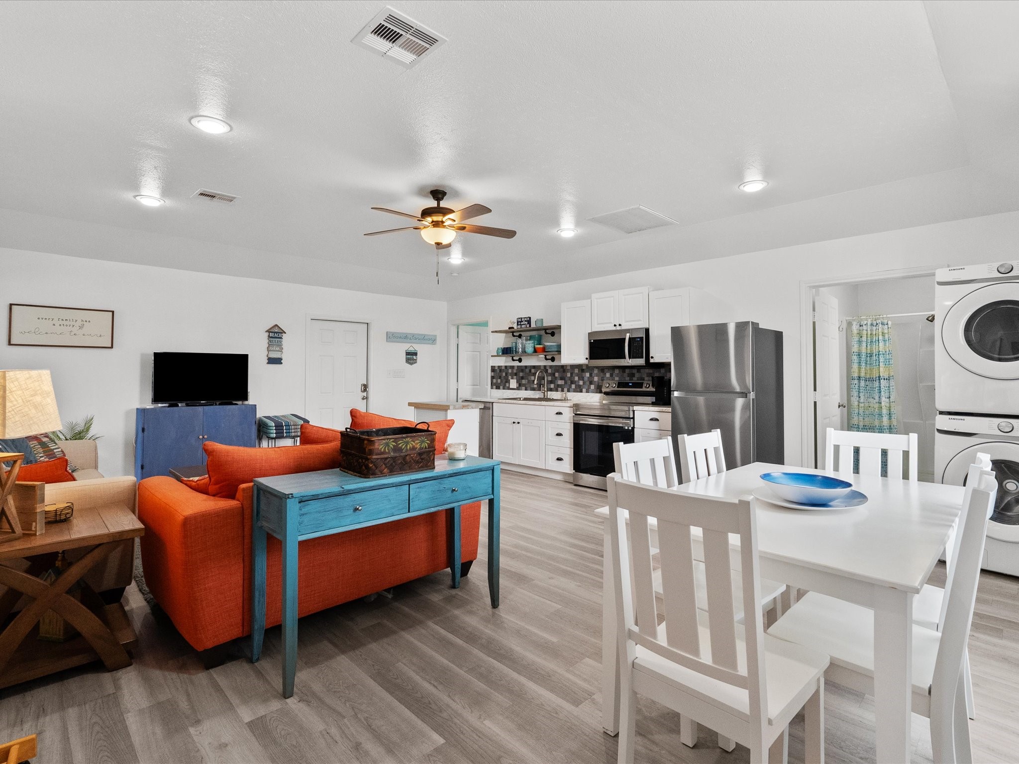 818 Surf Drive Surfside Beach, TX 77541 - Photo 11 of 29 This open-concept living space features a modern kitchen with stainless steel appliances, a cozy sitting area with a vibrant orange sofa, and a dining area with a white table. The room is bright with stylish wood flooring and a ceiling fan, while a stacked washer and dryer are conveniently tucked in the corner.