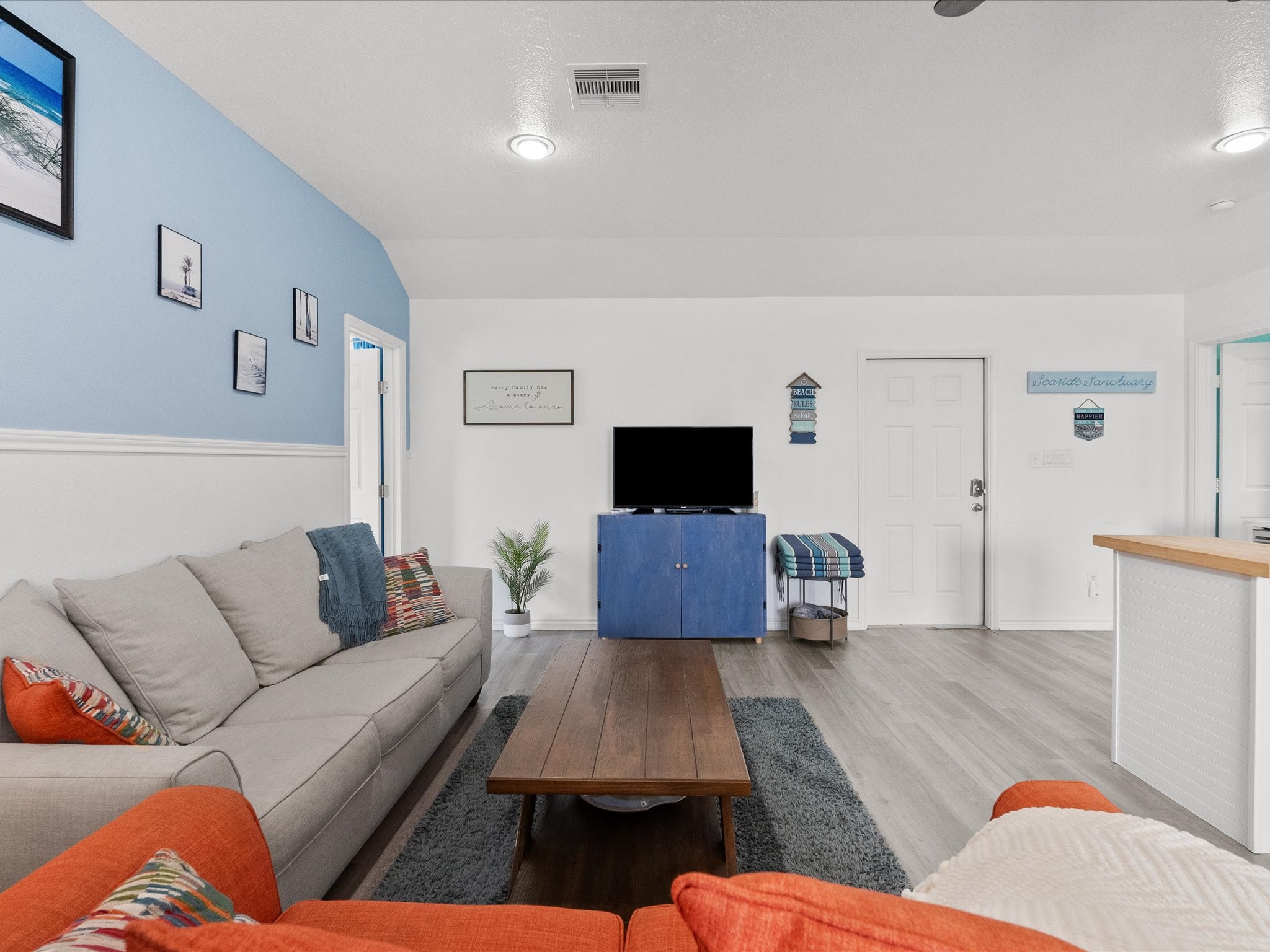 818 Surf Drive Surfside Beach, TX 77541 - Photo 13 of 29 This inviting living room features a cozy gray sofa and vibrant orange accents, complemented by a wooden coffee table. The room has a coastal theme with blue and white decor, a TV on a blue cabinet, and light wood flooring, creating a bright and welcoming atmosphere.