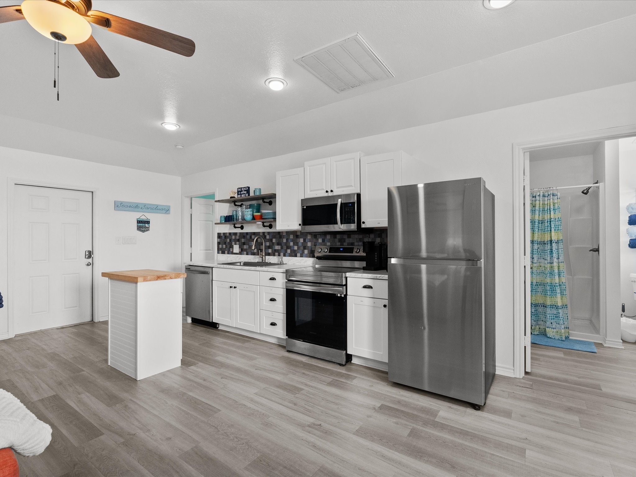 818 Surf Drive Surfside Beach, TX 77541 - Photo 14 of 29 Modern kitchen featuring stainless steel appliances, white cabinetry, and a sleek backsplash. The room is bright with recessed lighting and a ceiling fan. An island provides additional counter space, and a visible bathroom adds convenience.