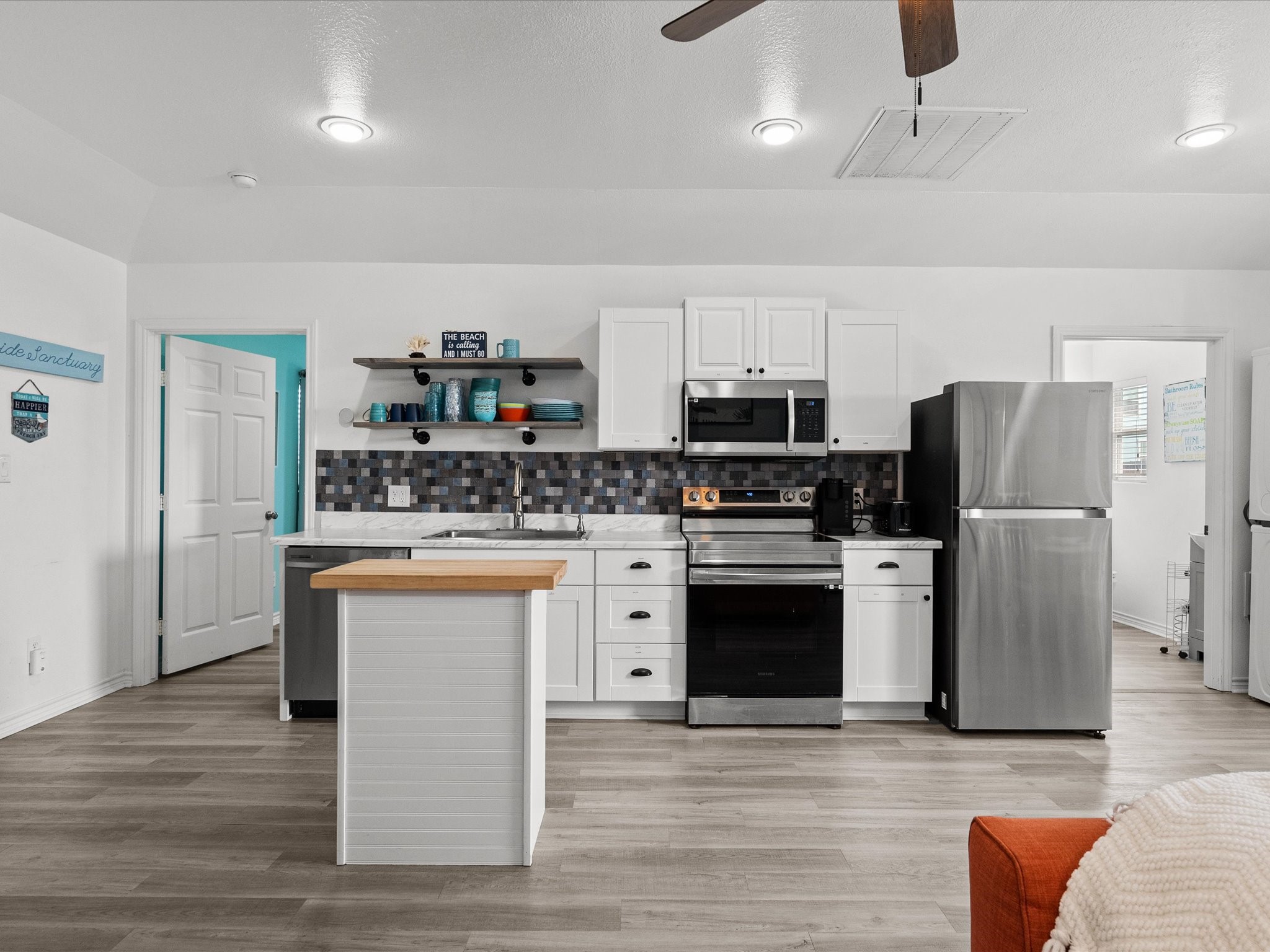 818 Surf Drive Surfside Beach, TX 77541 - Photo 15 of 29 This photo showcases a modern kitchen with sleek stainless steel appliances, white cabinetry, and a stylish tile backsplash. The space features a small island with a wooden countertop, open shelving, and light wood flooring, creating a bright and inviting atmosphere.