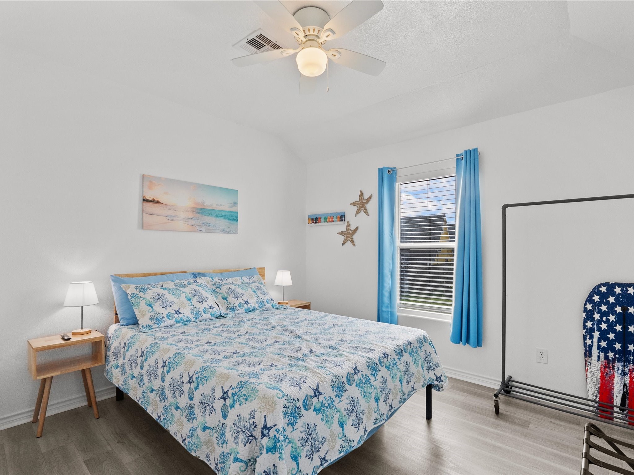 818 Surf Drive Surfside Beach, TX 77541 - Photo 18 of 29 This bedroom features a bright and airy design with white walls and a ceiling fan. It includes a comfortable bed with ocean-themed bedding, two bedside tables with lamps, and a window adorned with blue curtains. Coastal decor adds charm, and a clothing rack provides additional storage.