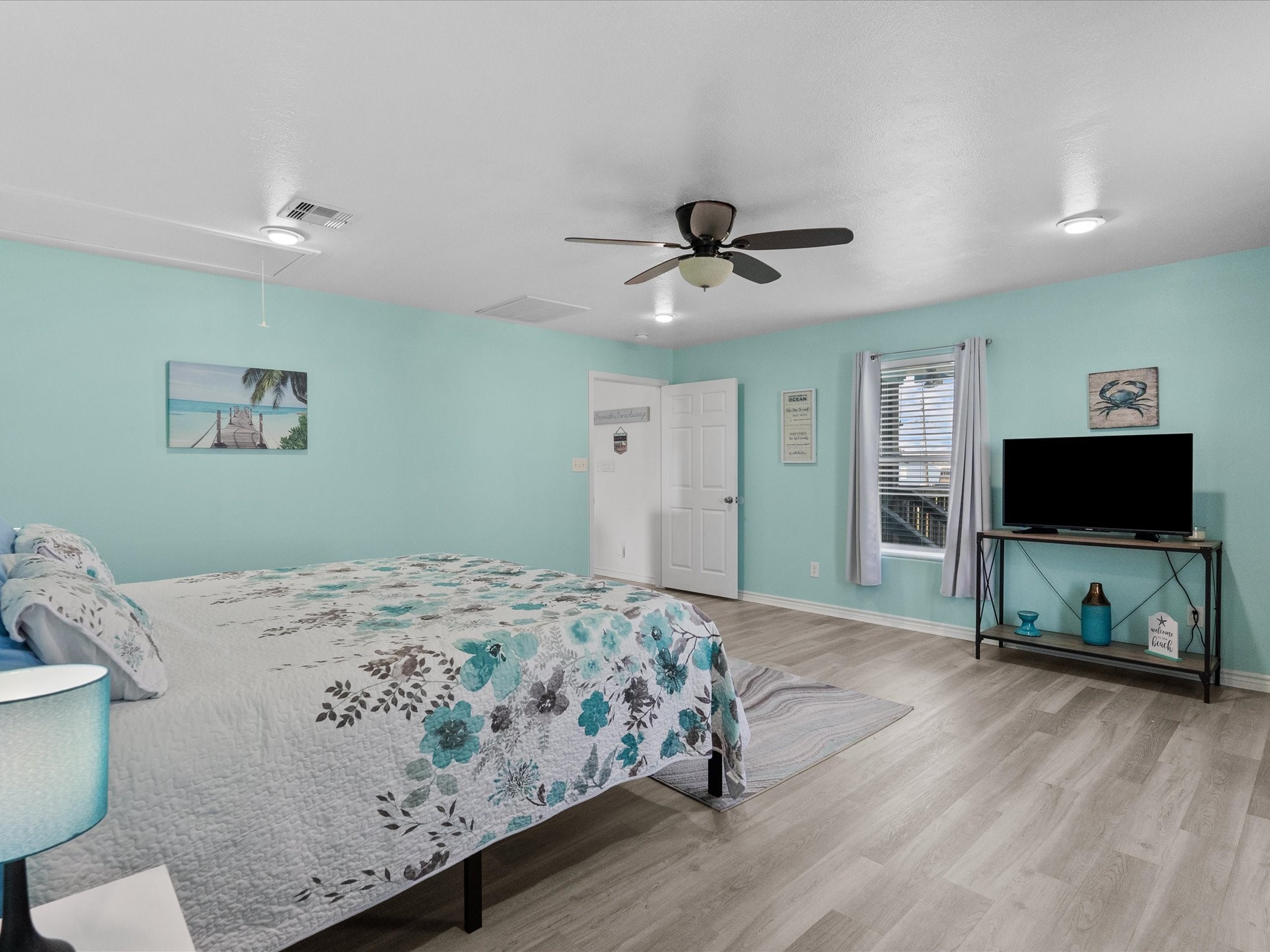 818 Surf Drive Surfside Beach, TX 77541 - Photo 21 of 29 Spacious bedroom with light teal walls, featuring a ceiling fan and wood-style flooring. It includes a comfortable bed with floral bedding, a TV on a stand, and a window with soft curtains, creating a calm, inviting atmosphere.