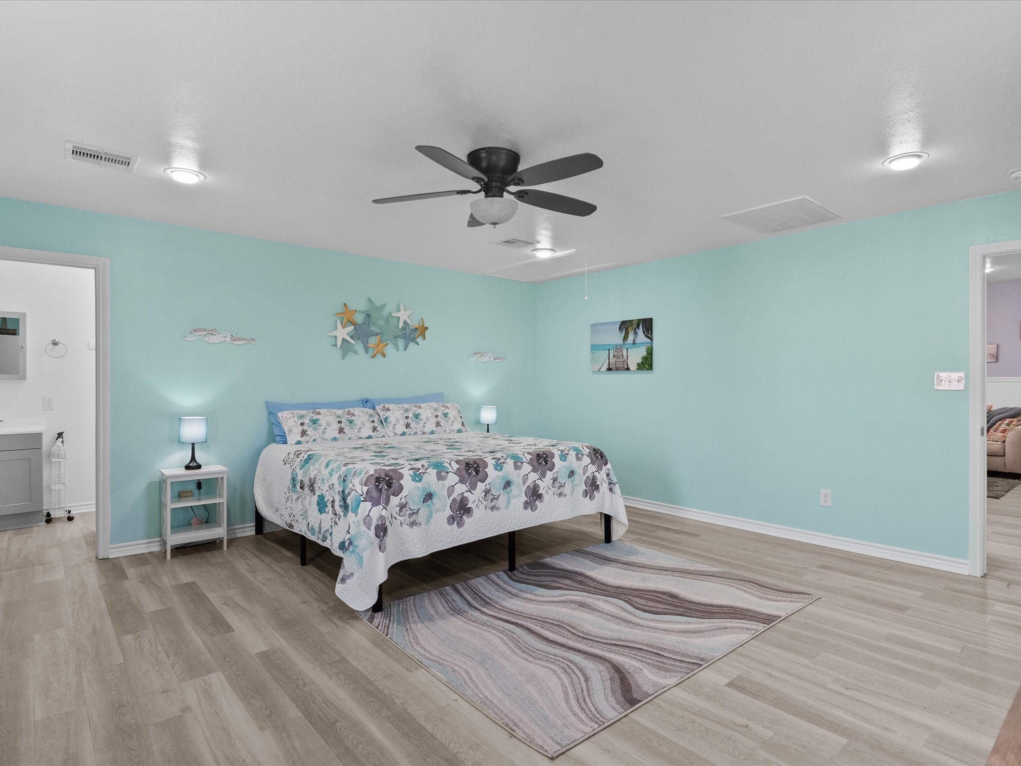 818 Surf Drive Surfside Beach, TX 77541 - Photo 22 of 29 This spacious bedroom features soothing aqua walls, light wood flooring, and a ceiling fan. It includes a comfortable bed with floral bedding, bedside tables with lamps, and decorative starfish wall art, creating a tranquil, beach-themed ambiance. A bathroom is accessible through an open doorway.