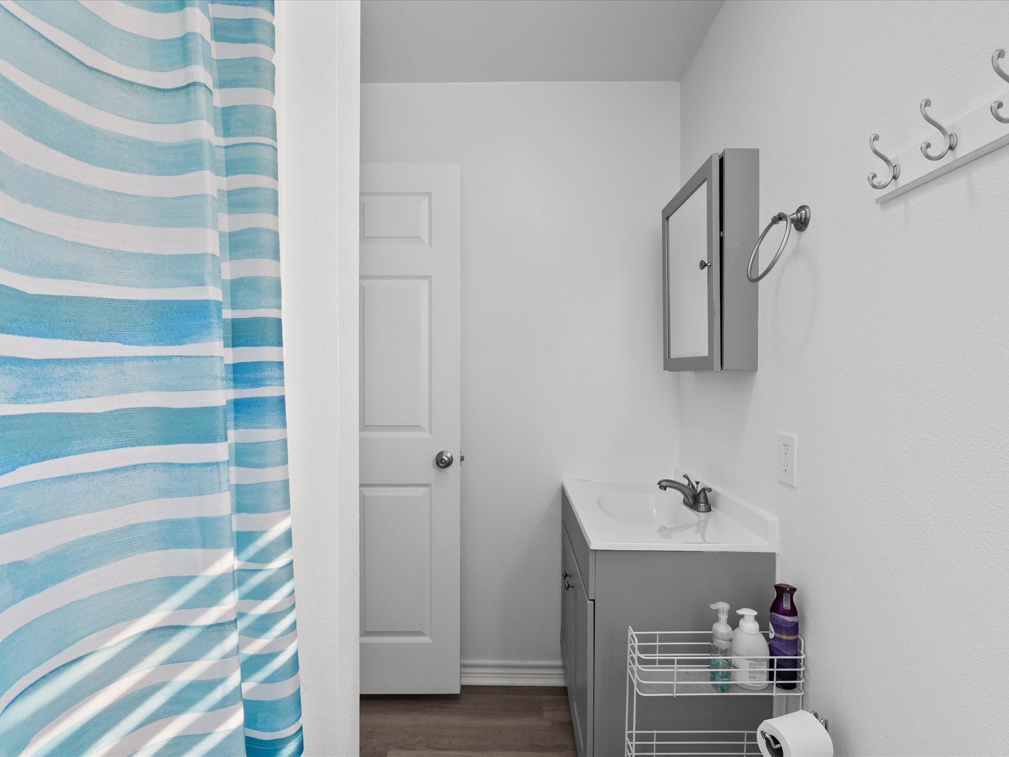 818 Surf Drive Surfside Beach, TX 77541 - Photo 23 of 29 Cozy bathroom with a modern aesthetic featuring a sleek gray vanity, wall-mounted mirror, and a vibrant blue-striped shower curtain. Hooks on the wall provide additional storage, enhancing functionality.