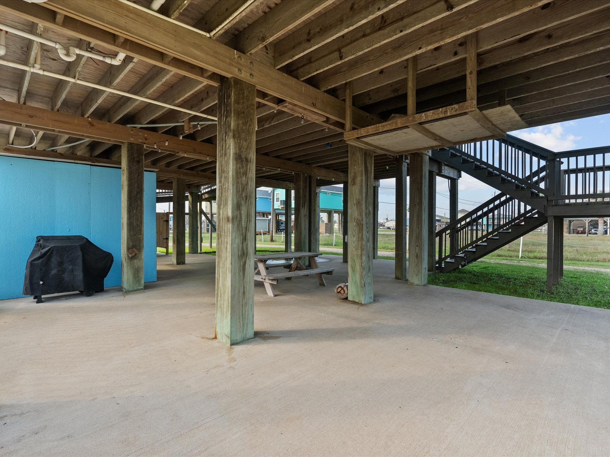 818 Surf Drive Surfside Beach, TX 77541 - Photo 26 of 29 This photo shows a covered outdoor space beneath a raised structure, featuring wooden beams and a staircase. There's a concrete floor, a picnic table, and a grill, ideal for outdoor gatherings.
