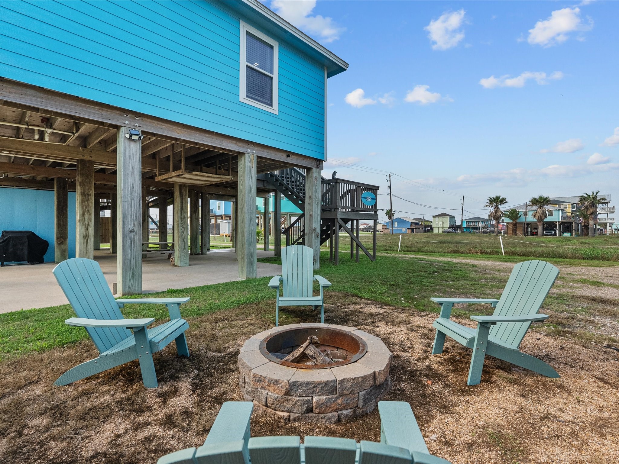 818 Surf Drive Surfside Beach, TX 77541 - Photo 28 of 29 This photo features a charming coastal home elevated on stilts, with a vibrant blue exterior. The outdoor area includes a cozy fire pit surrounded by Adirondack chairs, perfect for relaxing and enjoying the open, grassy surroundings.