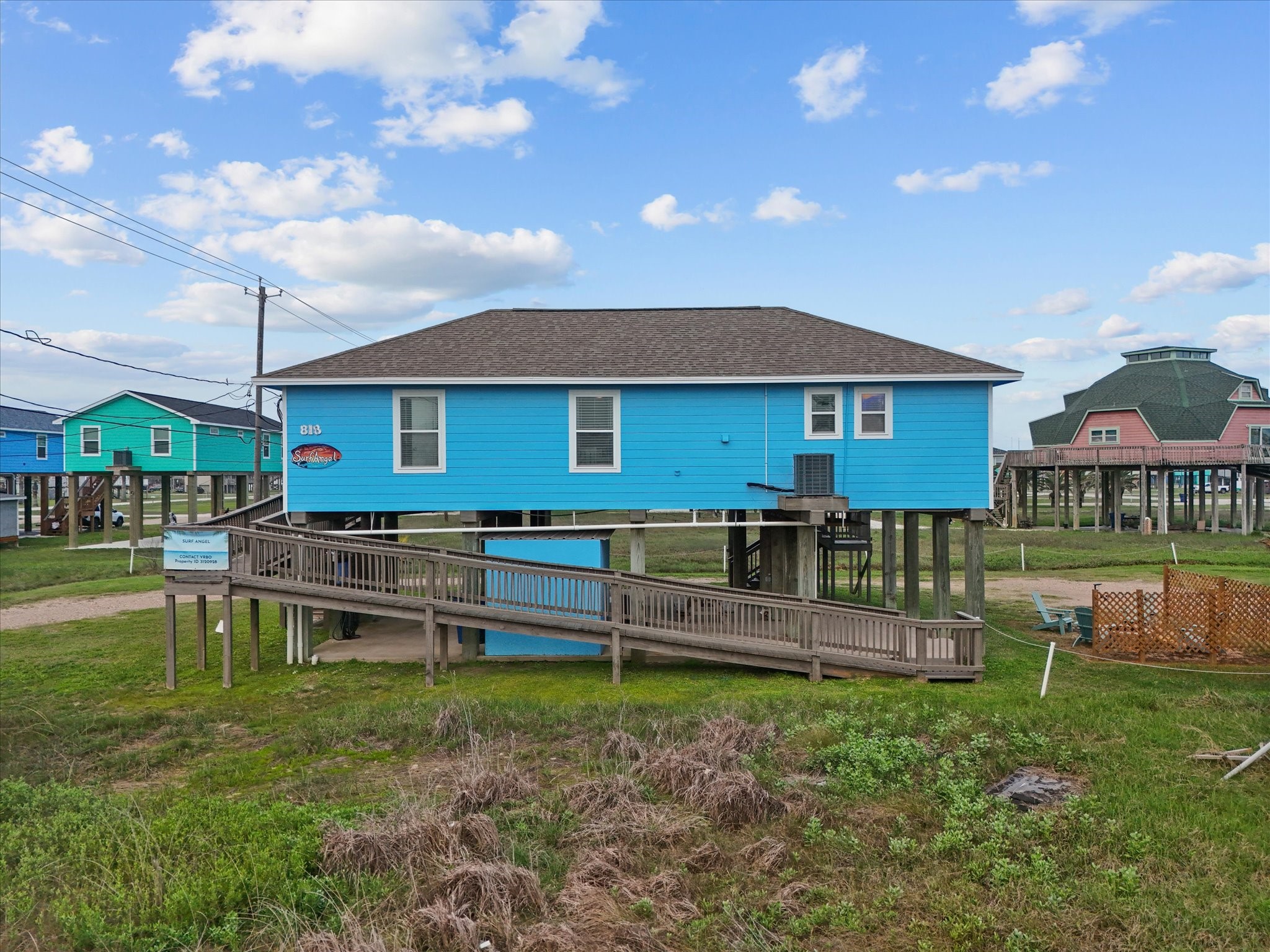 818 Surf Drive Surfside Beach, TX 77541 - Photo 5 of 29 Stilted beach house with vibrant blue exterior, featuring a ramp entrance in addition to the stairs and ample space underneath. Located in a coastal area with neighboring elevated homes.