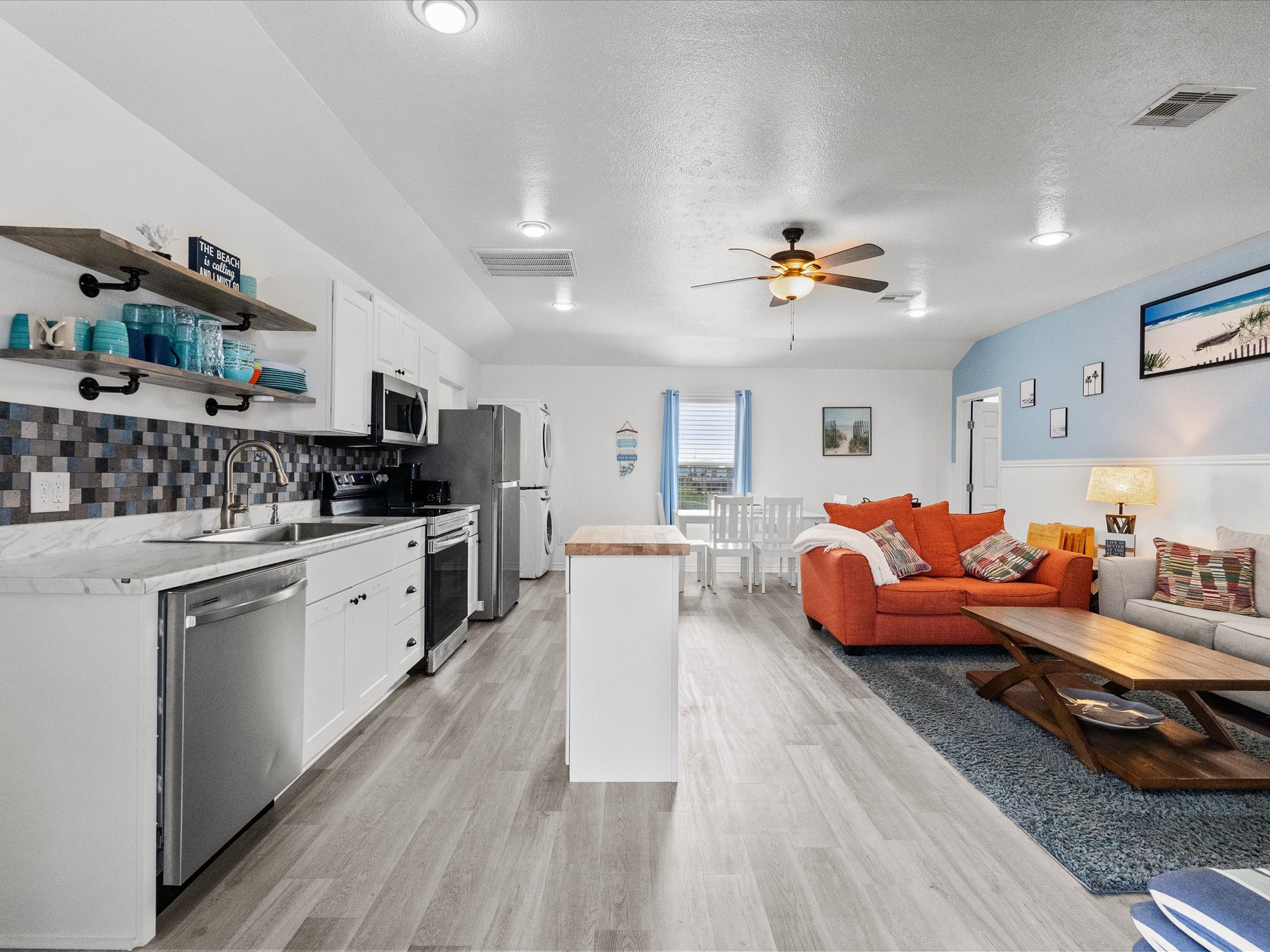 818 Surf Drive Surfside Beach, TX 77541 - Photo 8 of 29 This photo showcases a modern, open-concept kitchen and living area. The kitchen features white cabinets, stainless steel appliances, and a stylish backsplash. The living space includes a cozy orange sofa, a ceiling fan, and a dining area with natural light. The design is bright and inviting, perfect for entertaining.