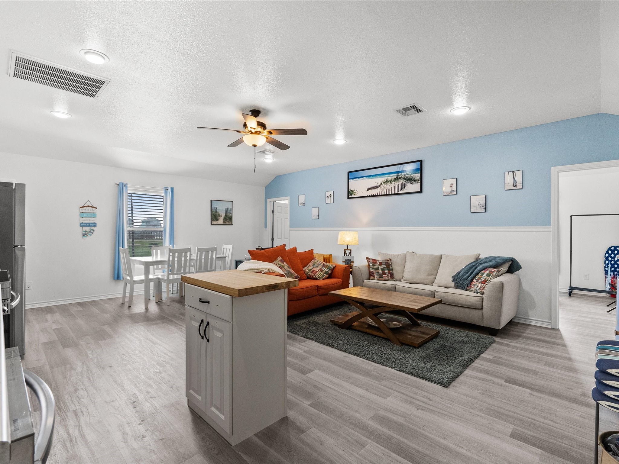 818 Surf Drive Surfside Beach, TX 77541 - Photo 9 of 29 This photo showcases a bright, open-concept living area with modern flooring and a cozy seating arrangement. The space features a light blue accent wall, ceiling fan, and ample natural light. A dining area with a white table and chairs is adjacent to the kitchen, which includes a central island for added functionality.