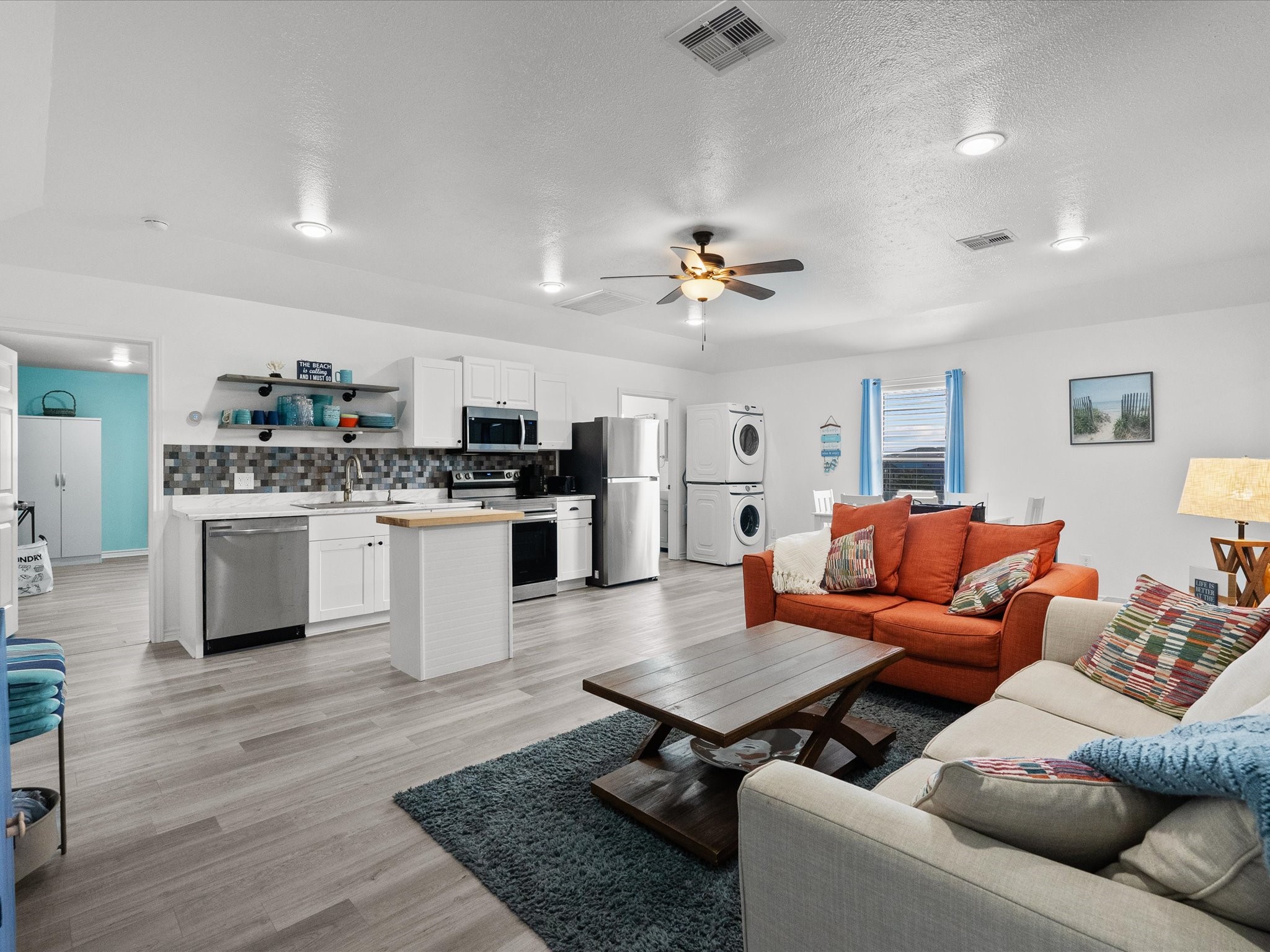 818 Surf Drive Surfside Beach, TX 77541 - Photo 10 of 29 This photo showcases a spacious, open-concept living area with modern kitchen amenities, including stainless steel appliances and a central island. The space is bright with recessed lighting, a ceiling fan, and features a cozy seating area with colorful decor. A washer and dryer are conveniently located in the corner, adding to the functionality.