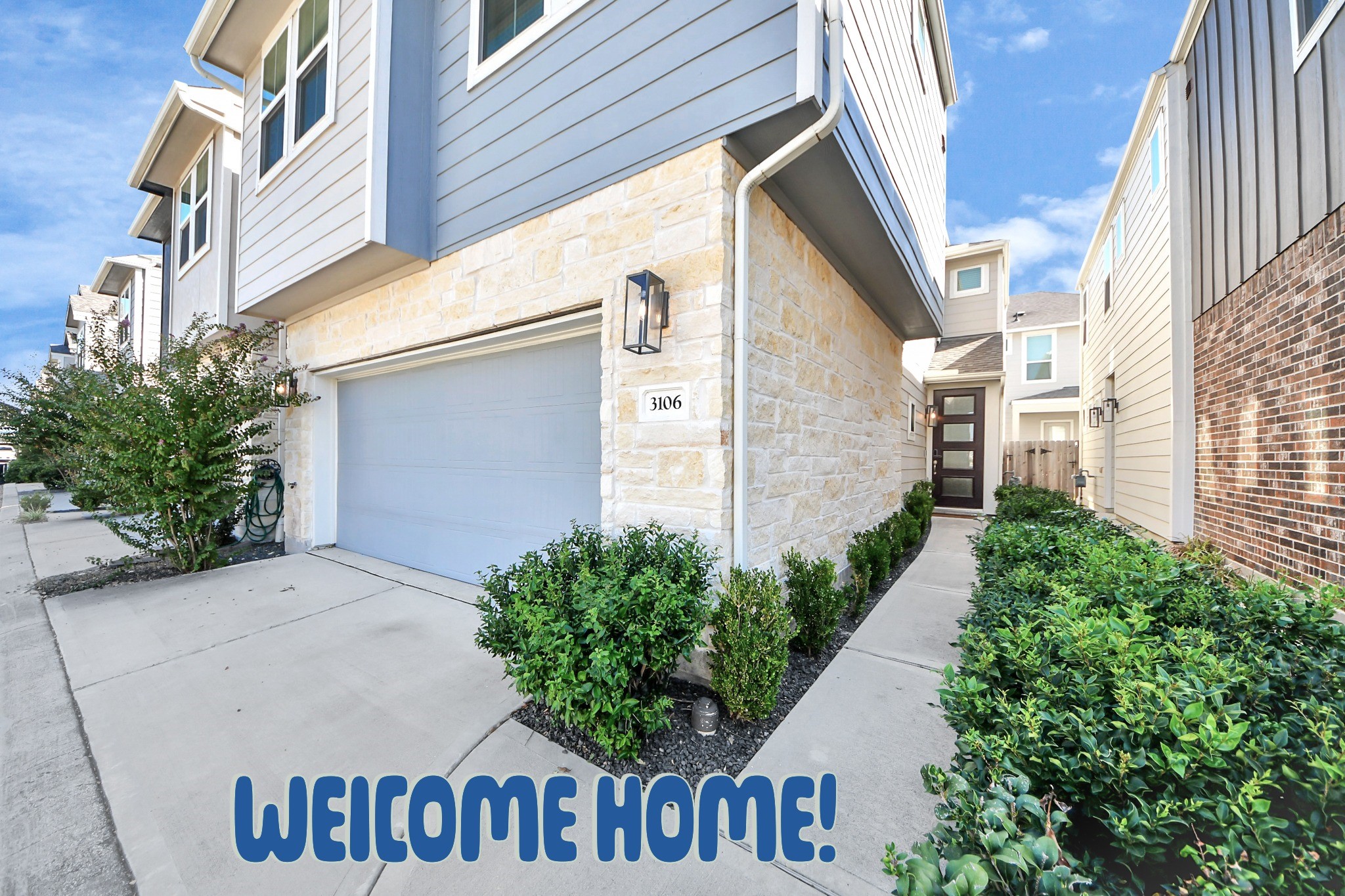 Modern home with stone and siding exterior, featuring a two-car garage. The entrance pathway is lined with manicured shrubs, leading to a welcoming front door.
