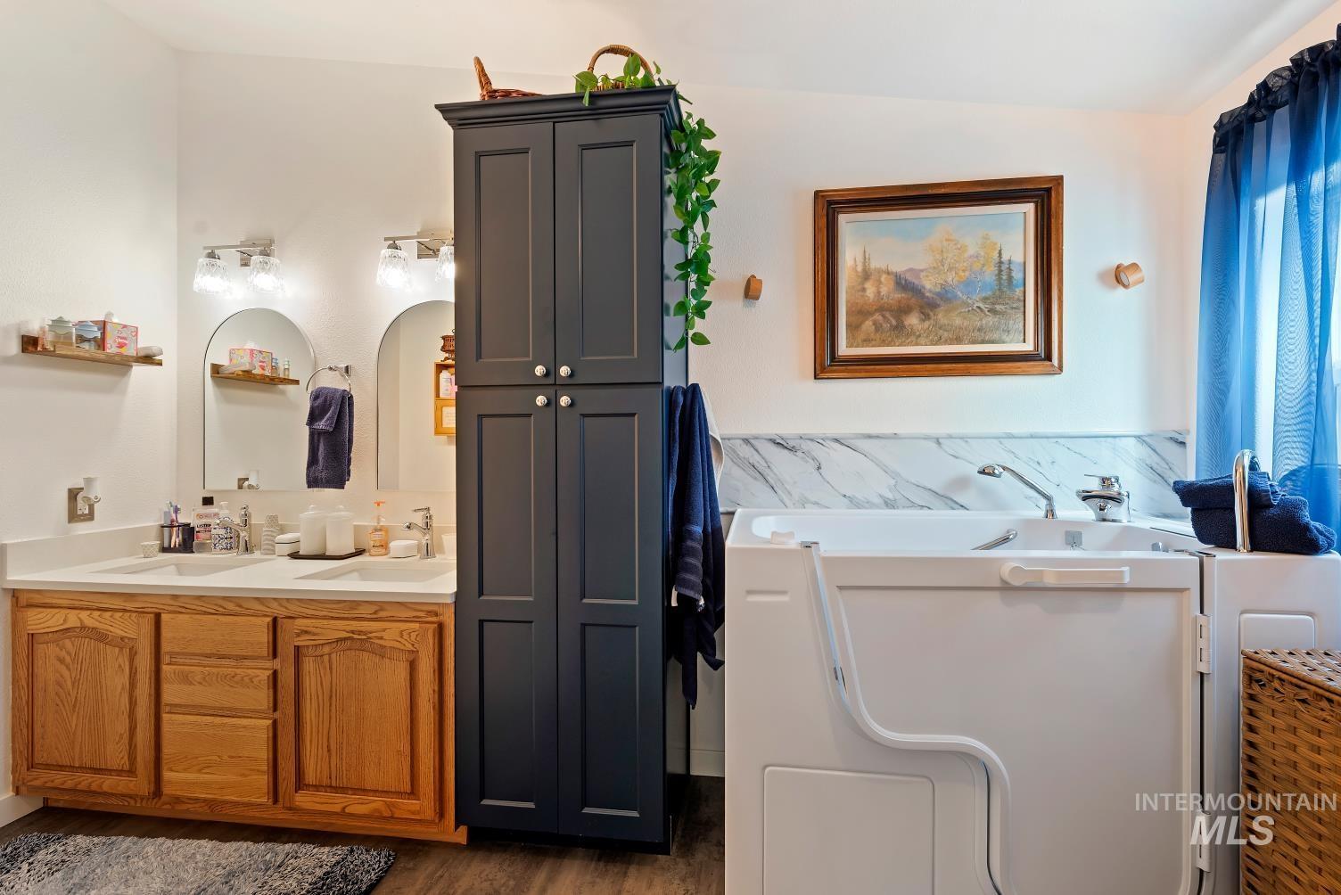 19152 Marsing Road Caldwell, ID 83607 - Photo 28 of 47 Bathroom featuring a walk-in tub, a dual vanity with light wood cabinetry, a tall dark gray storage cabinet, a white countertop, and marble-finish wainscoting