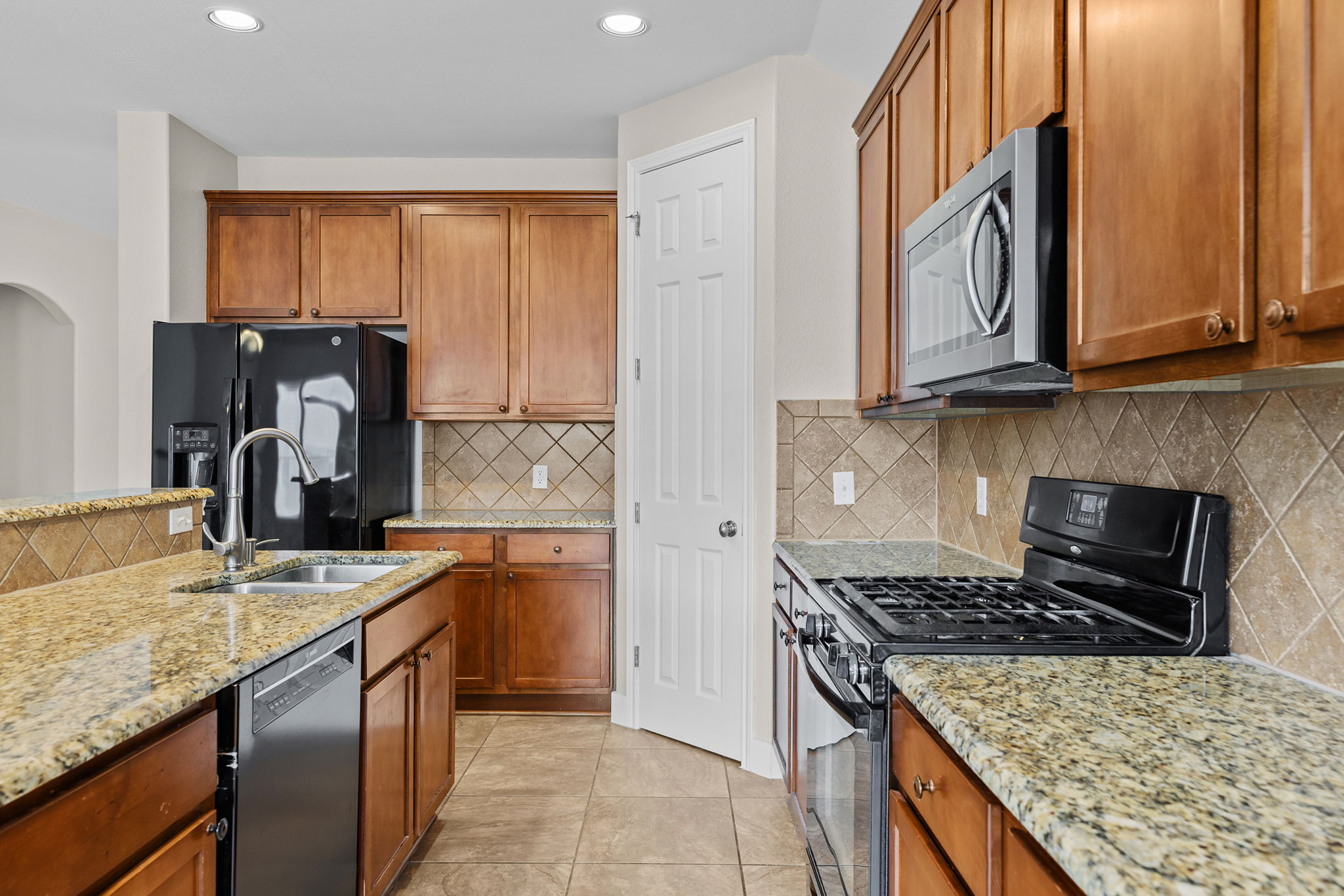 3013 Hawks Swoop Trail Pflugerville, TX 78660 - Photo 19 of 38 Kitchen featuring black appliances, wood finish cabinetry, backsplash, light stone counters, and light tile patterned flooring