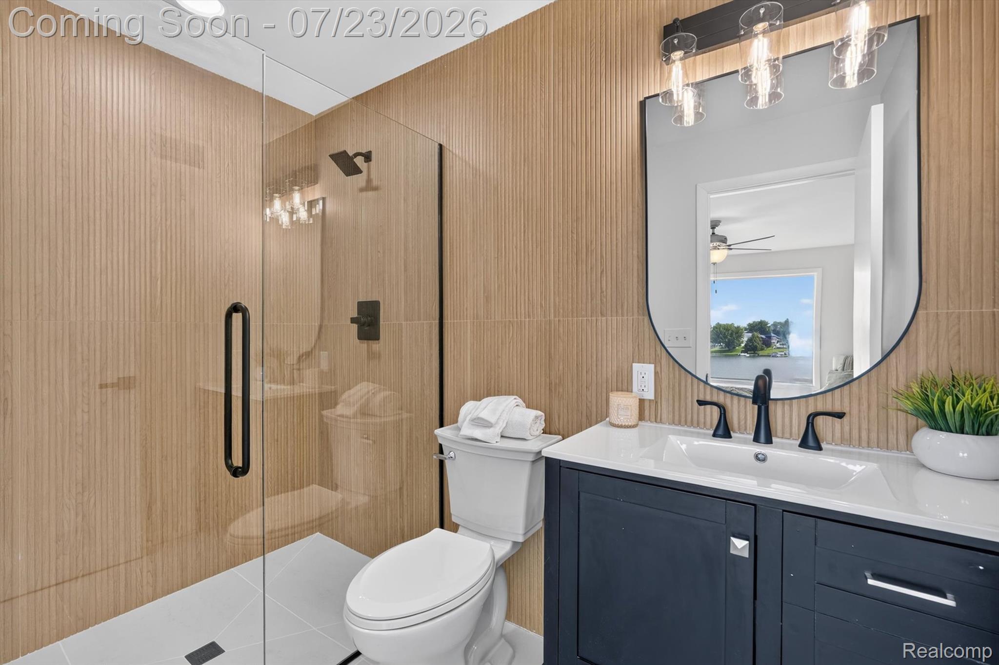 3429 Rolston Road Fenton, MI 48430 - Photo 18 of 62 Bathroom featuring a glass-enclosed shower with a black rain shower head, textured wood-finish wall tiling, a dark blue vanity with a white countertop and integrated sink, and a black-framed arched mirror with industrial-style lighting