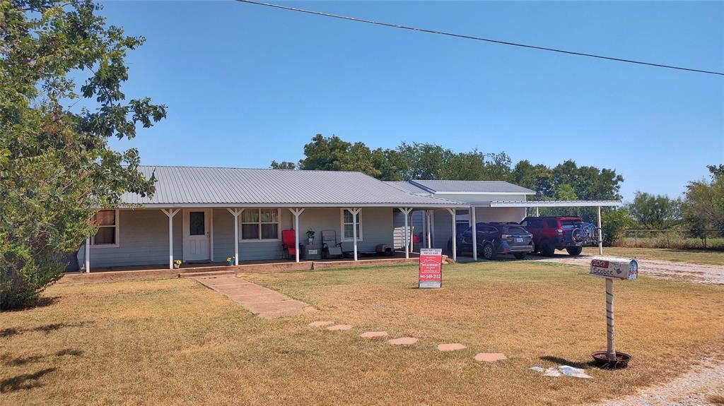 200 Scobee Road Olney, TX 76374 - Photo 1 of 1 Farmhouse-style home with a front lawn, an attached carport, a metal roof, driveway, and a porch
