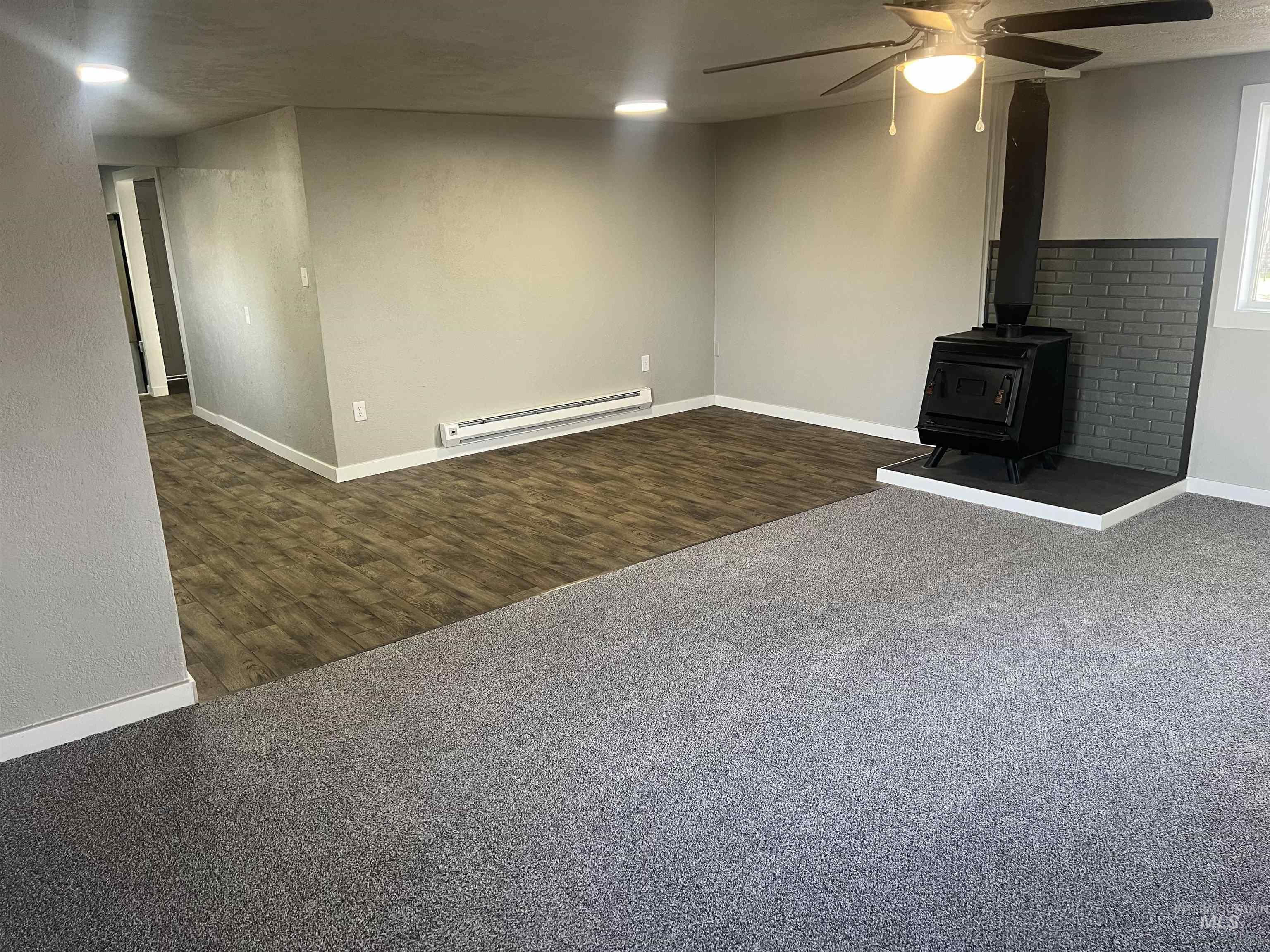 150 Roosevelt Street Mountain Home, ID 83647 - Photo 4 of 16 Unfurnished living room featuring a wood stove, dark colored carpet, a ceiling fan, a baseboard radiator, and dark wood-style flooring