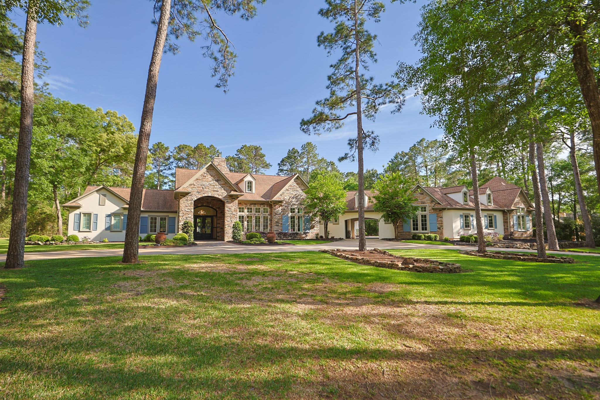 This stunning sprawling one story home features large windows, nestled among tall trees on a lush, well-manicured lawn. The architecture blends classic elegance with modern comfort, creating an inviting and picturesque setting.