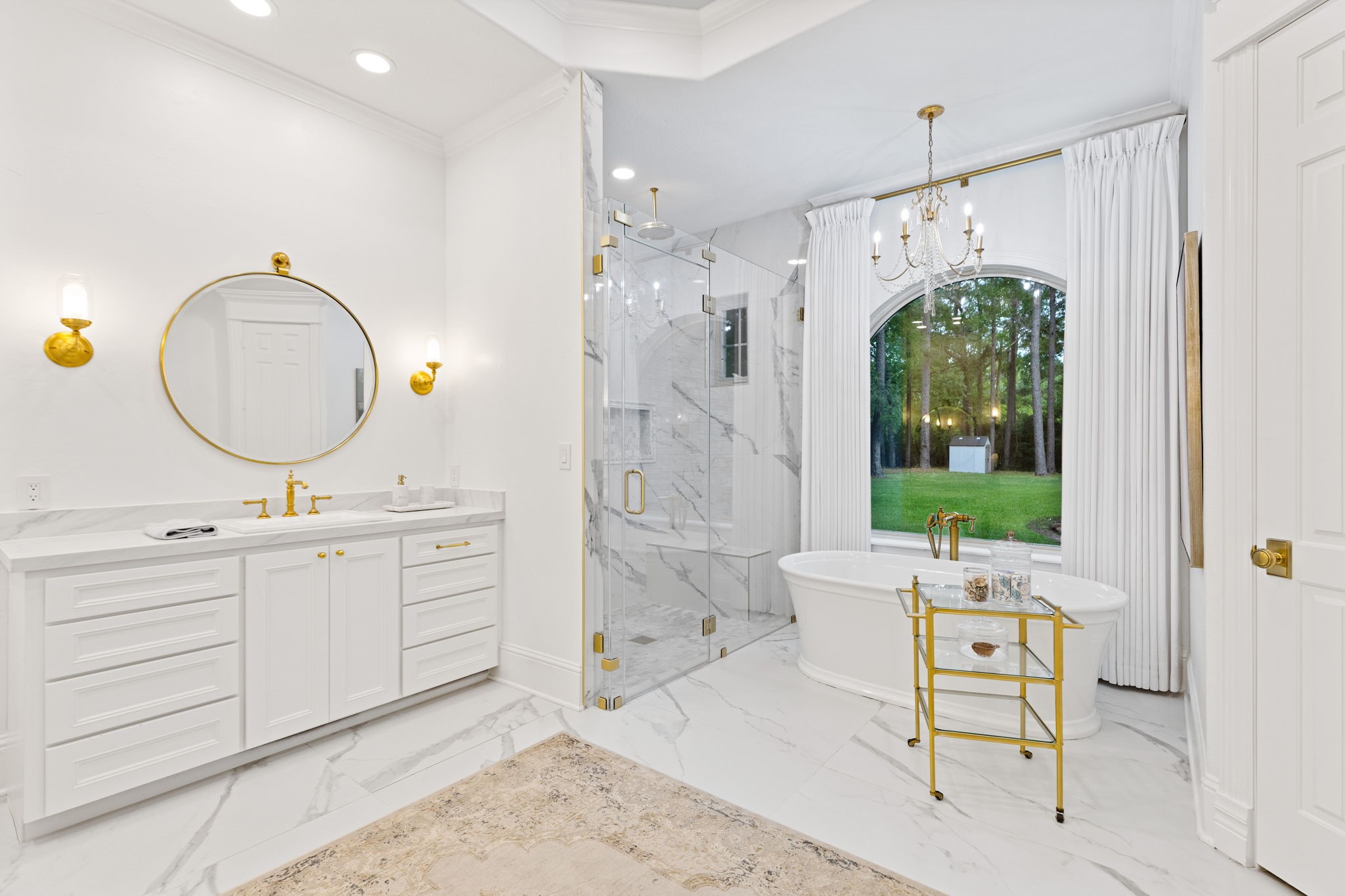 38223 Windy Ridge Trail Magnolia, TX 77355 - Photo 23 of 48 This bathroom features a luxurious design with a freestanding tub by a large window, a glass-enclosed marble shower, and double vanity with gold fixtures. Elegant lighting and a scenic outdoor view enhance the serene atmosphere. Simply gorgeous!