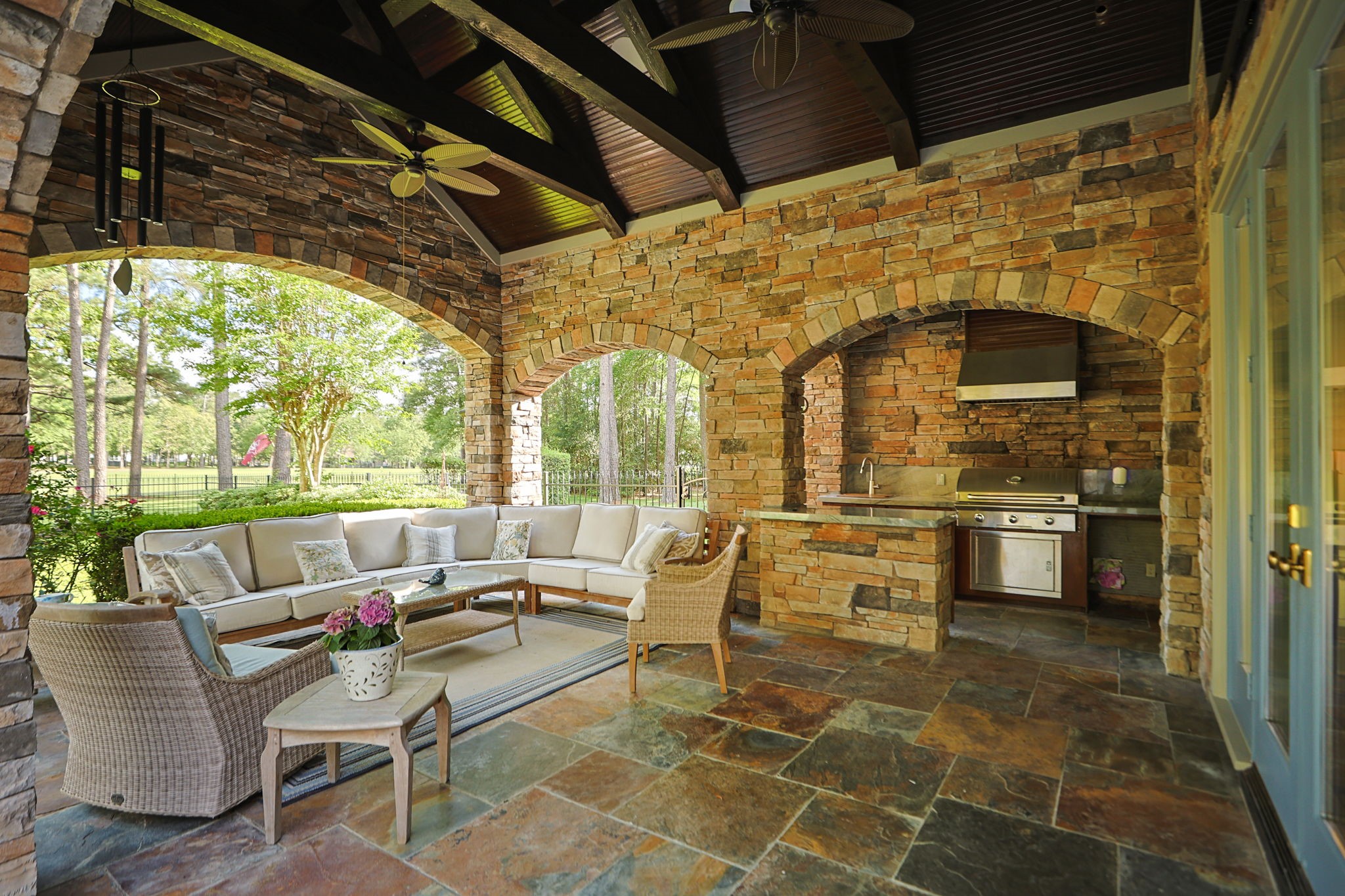 38223 Windy Ridge Trail Magnolia, TX 77355 - Photo 33 of 48 This photo showcases a beautiful outdoor living space with a rustic stone design, featuring a cozy seating area, built-in grill, ceiling fans, and beaded board ceiling. The arched openings provide a scenic view of the lush greenery outside. Perfect for entertaining and relaxing.