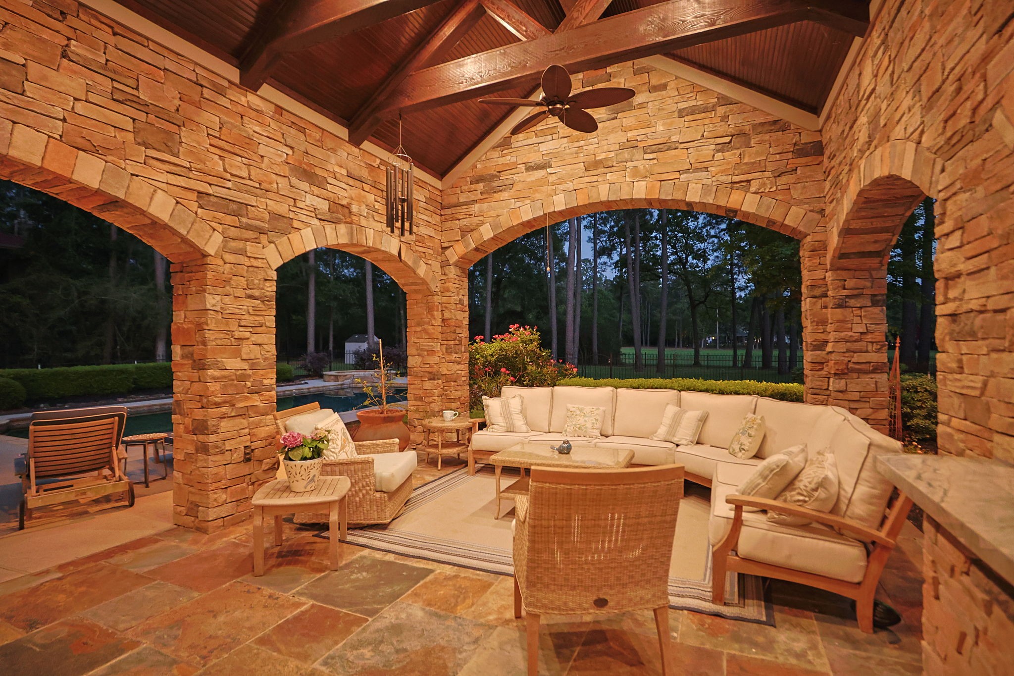 38223 Windy Ridge Trail Magnolia, TX 77355 - Photo 34 of 48 This sunset photo of the inviting outdoor living space features a vaulted wooden ceiling with fans. Surrounded by elegant stone arches, it offers a serene view of a lush, wooded area, perfect for relaxation or entertaining.