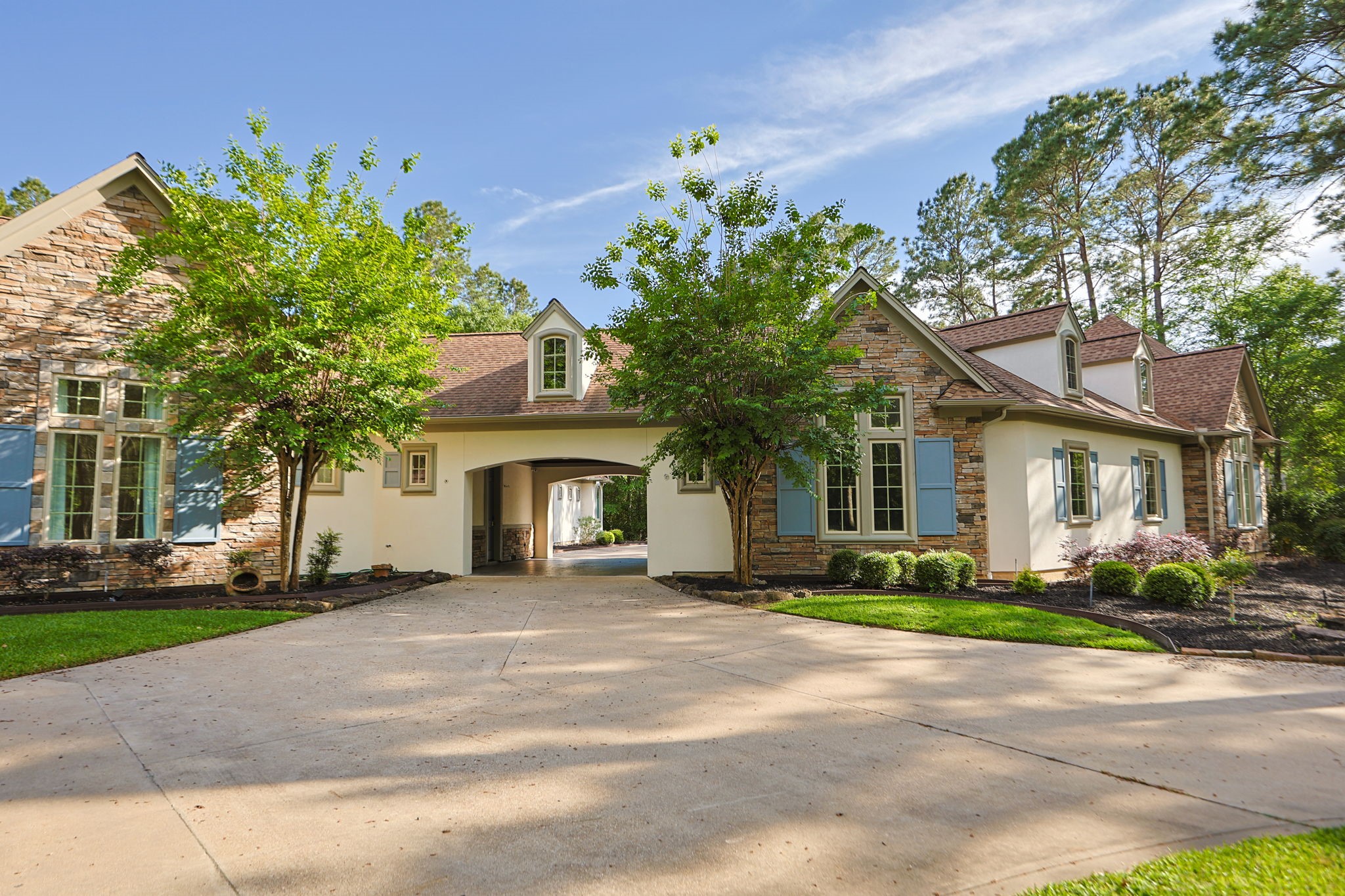 38223 Windy Ridge Trail Magnolia, TX 77355 - Photo 5 of 48 This photo showcases a charming, single-story home with stone and stucco exterior, warm blue shutters, and a welcoming port-te-chere. It features a spacious driveway, and beautiful lush landscaping.