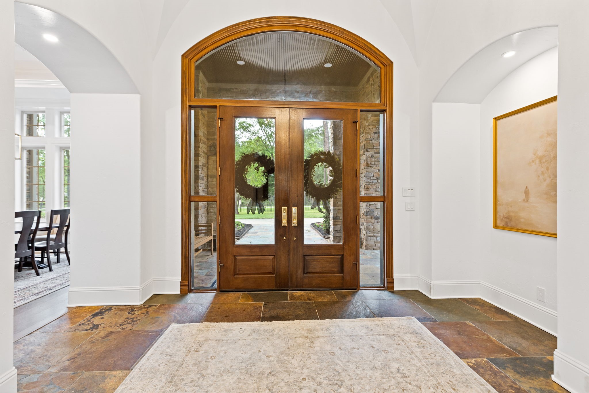 38223 Windy Ridge Trail Magnolia, TX 77355 - Photo 8 of 48 This photo showcases a spacious, elegant entryway with a large beautiful wooden double door featuring glass panels. The floor is slate tile with a warm, earthy tone. The area is well-lit with recessed lighting, and a glimpse of a dining room with large windows is visible to the left.