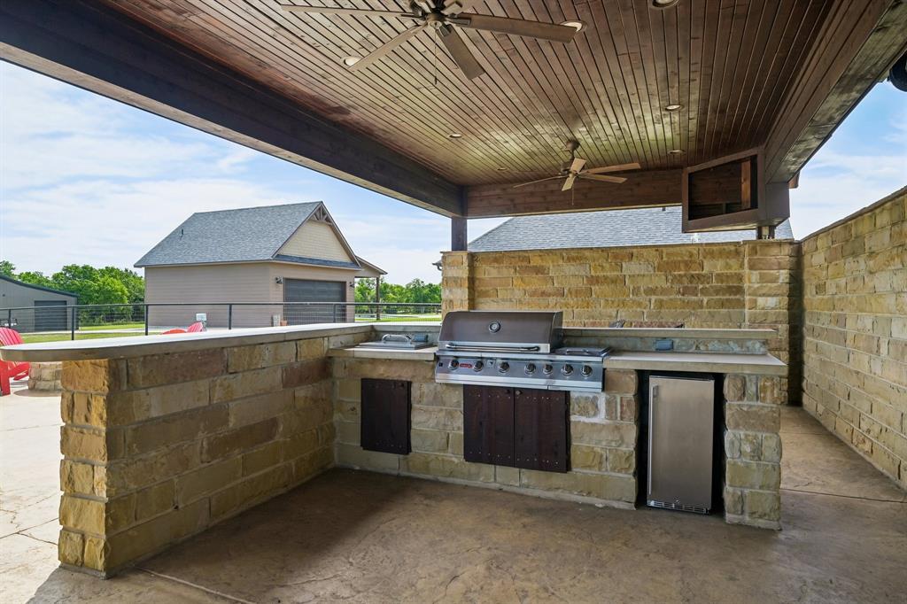 2085 Switzer Road Sanger, TX 76266 - Photo 32 of 39 Covered outdoor kitchen featuring a built-in grill, integrated beverage refrigerator, stone-finish cabinetry, and a wood-slat ceiling with two ceiling fans