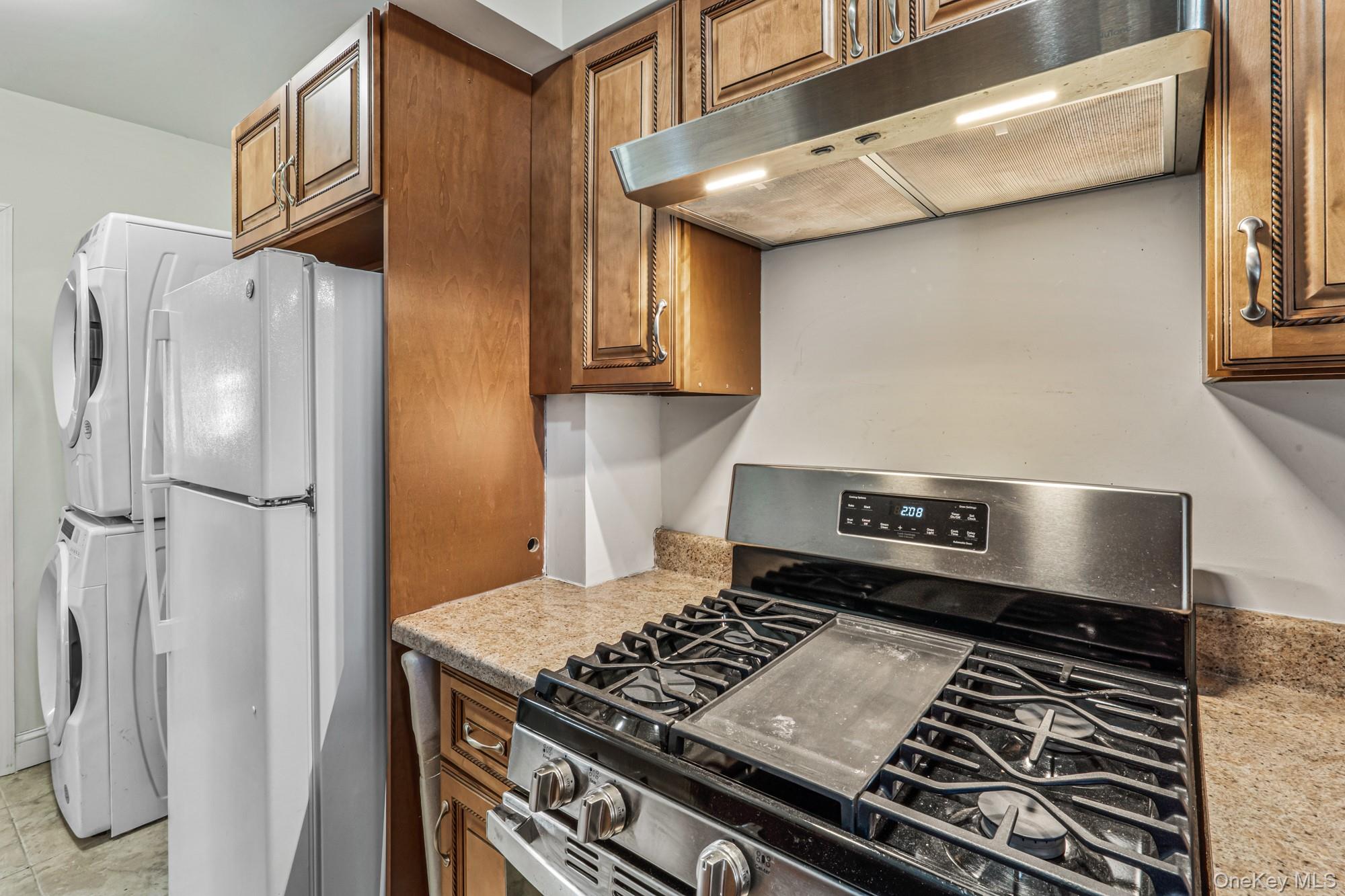 61-12 Main Street Queens, NY 11367 - Photo 20 of 35 Kitchen featuring stainless steel range with gas stovetop, under cabinet range hood, light stone countertops, brown cabinets, and freestanding refrigerator